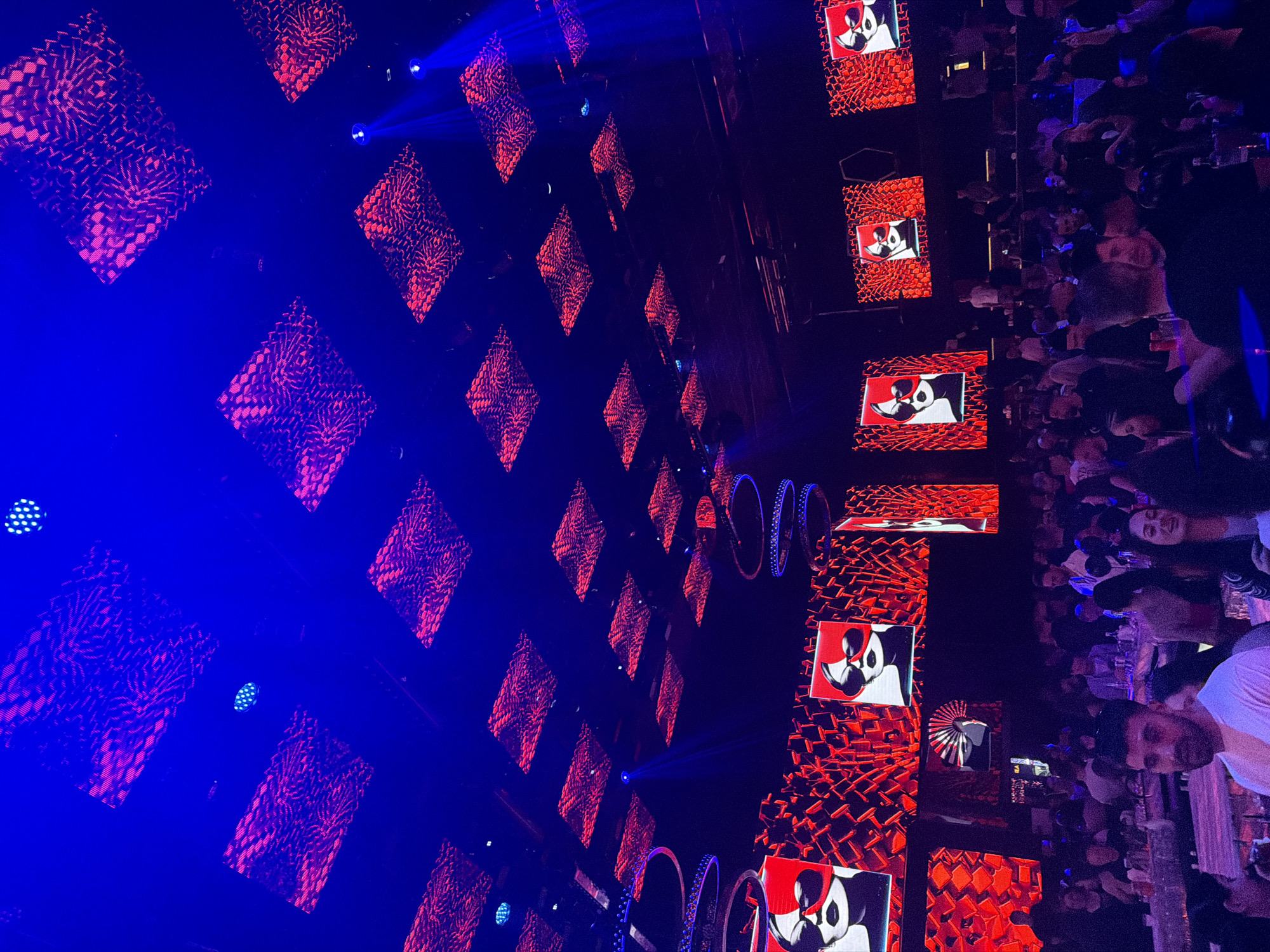The image shows a nighttime scene, likely indoors at a club or entertainment venue in an unknown city, judging by the crowd and stage setup. On the left, there's a wall-like structure with repeating diamond-shaped designs illuminated in red and blue light. This appears to be a stage backdrop or artistic installation. Blue spotlights are visible.
To the right, a crowd of people is visible, some looking towards the camera or the stage. Some of the crowd members have drinks. The stage backdrop has rectangular screens showing an animation of a black and white dog. There are also illuminated round decorative hoops.