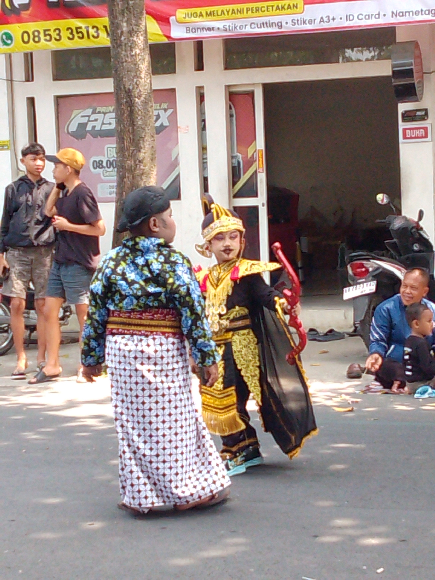 The image captures a street scene in Trenggalek, Indonesia, likely during a cultural event or procession. Two children are the central figures. One child, dressed in traditional attire with a black and blue floral patterned top and a white batik sarong, stands facing away from the camera. The other child, adorned in a golden and black costume with elaborate headdress and holding a red bow, is positioned in front of the first child, facing left.
In the background, two young men are visible standing near a building with signs. One man wears a black jacket, and the other wears a black t-shirt and shorts. To the right, a man in a blue jacket is seated on the ground, looking at a small child dressed in black. A motorcycle is parked in front of the building. The street appears to be paved, with some scattered leaves and shadows cast by overhead foliage. A banner with Indonesian text and a phone number is displayed on the left side of the frame. The environment suggests a sunny day in an urban setting.