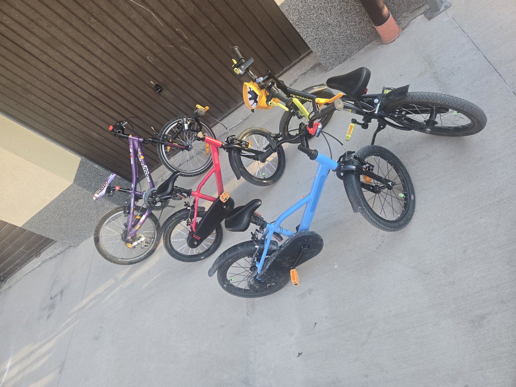 The image shows four children's bicycles parked in a row against a building. The bikes are various colors: purple, red, blue, and black with yellow accents. The building has a brown garage door and a section of grey stone siding. The ground is light grey concrete. There are no people visible. The location appears to be an outdoor area, possibly a driveway or patio, in an urban setting.