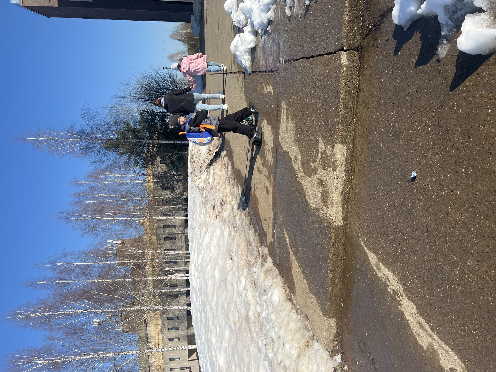 The photo shows a street scene in Марковский, Russia, likely in early spring. The weather is sunny with a clear blue sky. Patches of melting snow are visible along the side of the road and adjacent to a concrete curb. A row of bare birch trees lines the background.
Three people are walking on the sidewalk. The person closest to the camera is a child wearing a dark jacket, pants, and shoes, with a backpack. Further back, there are two more people: one wearing a dark jacket and light-colored pants, and another wearing a light pink jacket and light-colored pants. The people are walking on a wet, asphalt road. A discarded piece of trash is on the ground. A multi-story building is partially visible in the background.