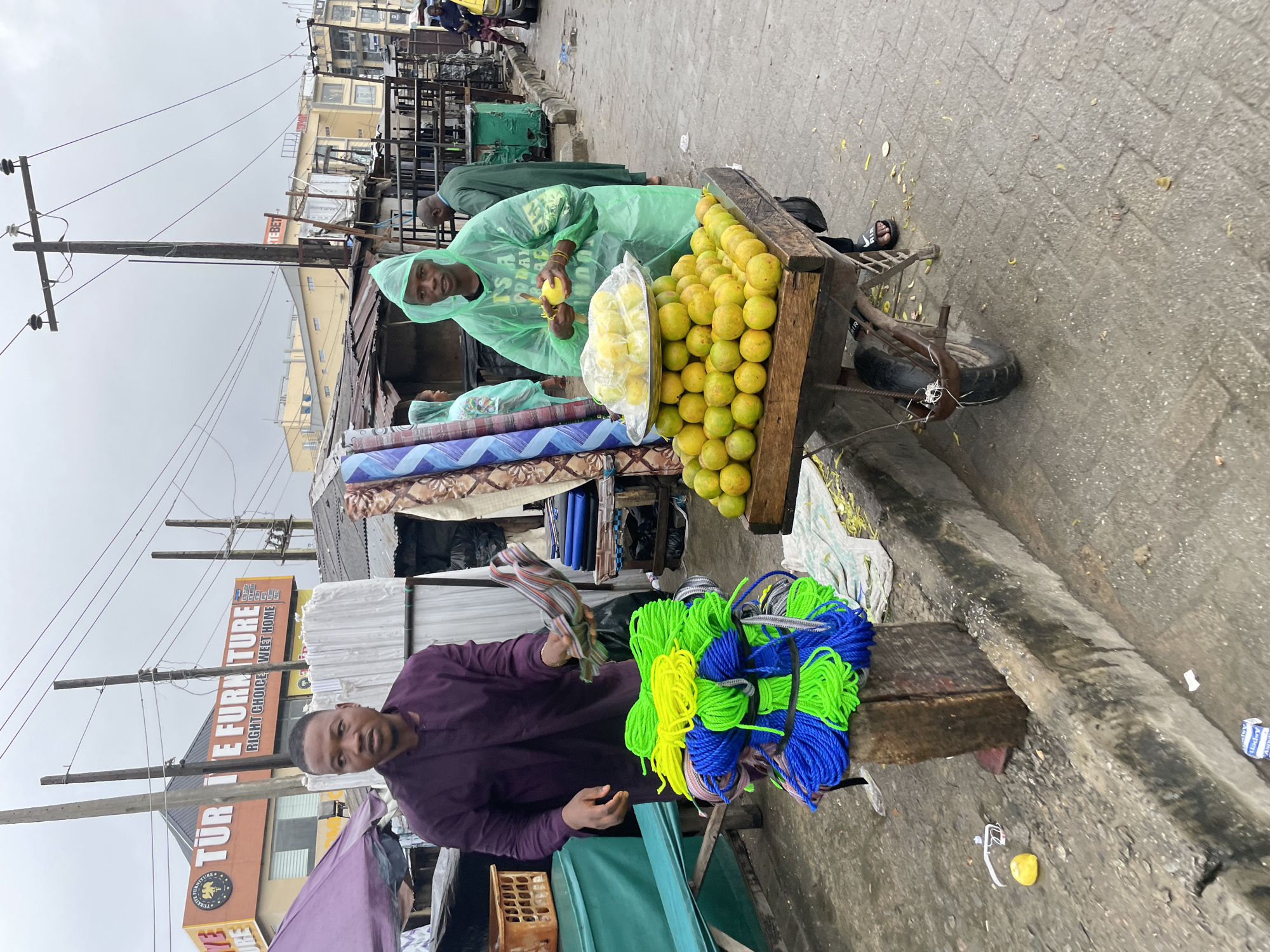 The image depicts a street scene in Lagos, Nigeria, likely a market area. In the foreground, a man in a purple traditional outfit stands next to a stack of brightly colored ropes. To his right, another man, wearing a green raincoat, is attending to a cart filled with oranges. The cart is positioned on a paved sidewalk next to a curb. In the background, various stalls and buildings are visible, including a shop with a prominent sign for "TÜR TE FURNITURE." The sky is overcast, suggesting cloudy weather. The overall environment appears to be a busy urban commercial district.