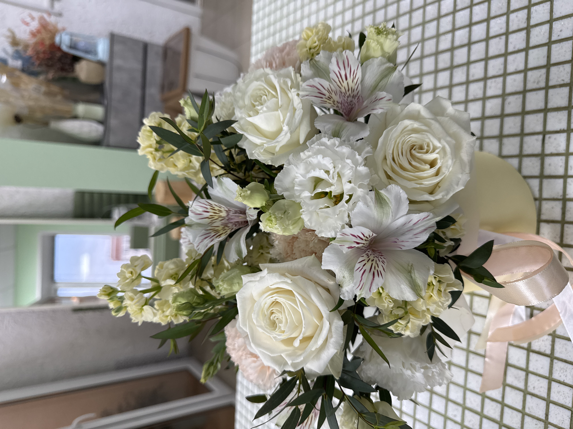 The image displays a detailed, well-lit close-up of a fresh floral bouquet presented in a light yellow/cream cylindrical vase, situated on a table covered with a white tablecloth featuring a light green/grey grid pattern. The bouquet primarily consists of various white flowers, including prominent white roses with tightly wound petals, white alstroemeria characterized by striped purple-burgundy markings on their inner petals, ruffled white Eustoma (Lisianthus), and white carnations. Interspersed among these are light yellow/cream flowers (resembling stock) and delicate light pink/peach colored smaller flowers. Dark green foliage, consistent with eucalyptus or similar leaves, provides contrast and fills the arrangement. Two ribbons, one pale pink and one cream/pale yellow, are tied around the vase, cascading downwards on the left side.
The background, largely out of focus, reveals an indoor setting, likely a retail space such as a flower shop, located in the city of Izhevsk, Russia. To the left, a blurred reflection in a large mirror or window frame shows a muted brown/peach tone and white vertical structures. Central to the background is a window or doorway offering a clear view to the outside, depicting a bright blue sky and the distant outlines of buildings, some with reddish rooftops, under daylight conditions. To the right of this opening, a light green wall or panel is visible. Further right, a blurred shelving unit or console table holds an assortment of decorative objects, including various white, light green, and beige vases, as well as dried floral arrangements in hues of natural brown, blue, and reddish-orange. A light blue decorative lantern is also present on this unit. No individuals are visible in the scene.