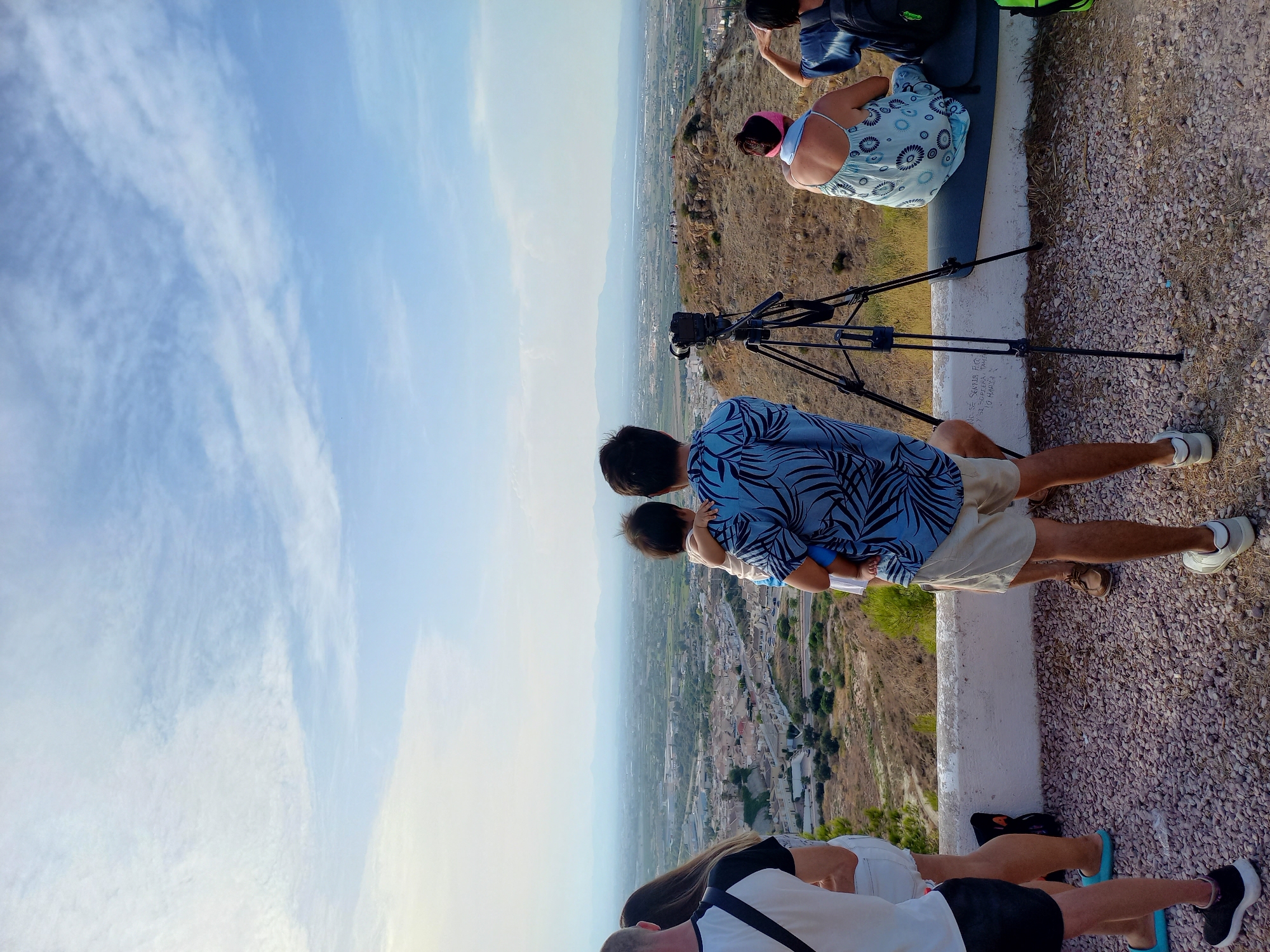 The image depicts a group of people at an elevated viewpoint overlooking the urban landscape of Ciudad Quesada, Spain. In the foreground, several individuals are visible from the waist down, including a man holding a baby, another man in a blue patterned shirt, and two women. The man holding the baby is wearing tan shorts, and the other man is wearing a blue and black patterned shirt. One woman is wearing white shorts, and the other is wearing a light blue patterned dress. A camera on a tripod is positioned in the middle ground, facing the panoramic view. The background reveals a sprawling town with numerous buildings, nestled amongst green fields and mountains in the distance. The sky is partly cloudy with a clear blue expanse. The ground in the foreground is covered with small, light-colored pebbles.