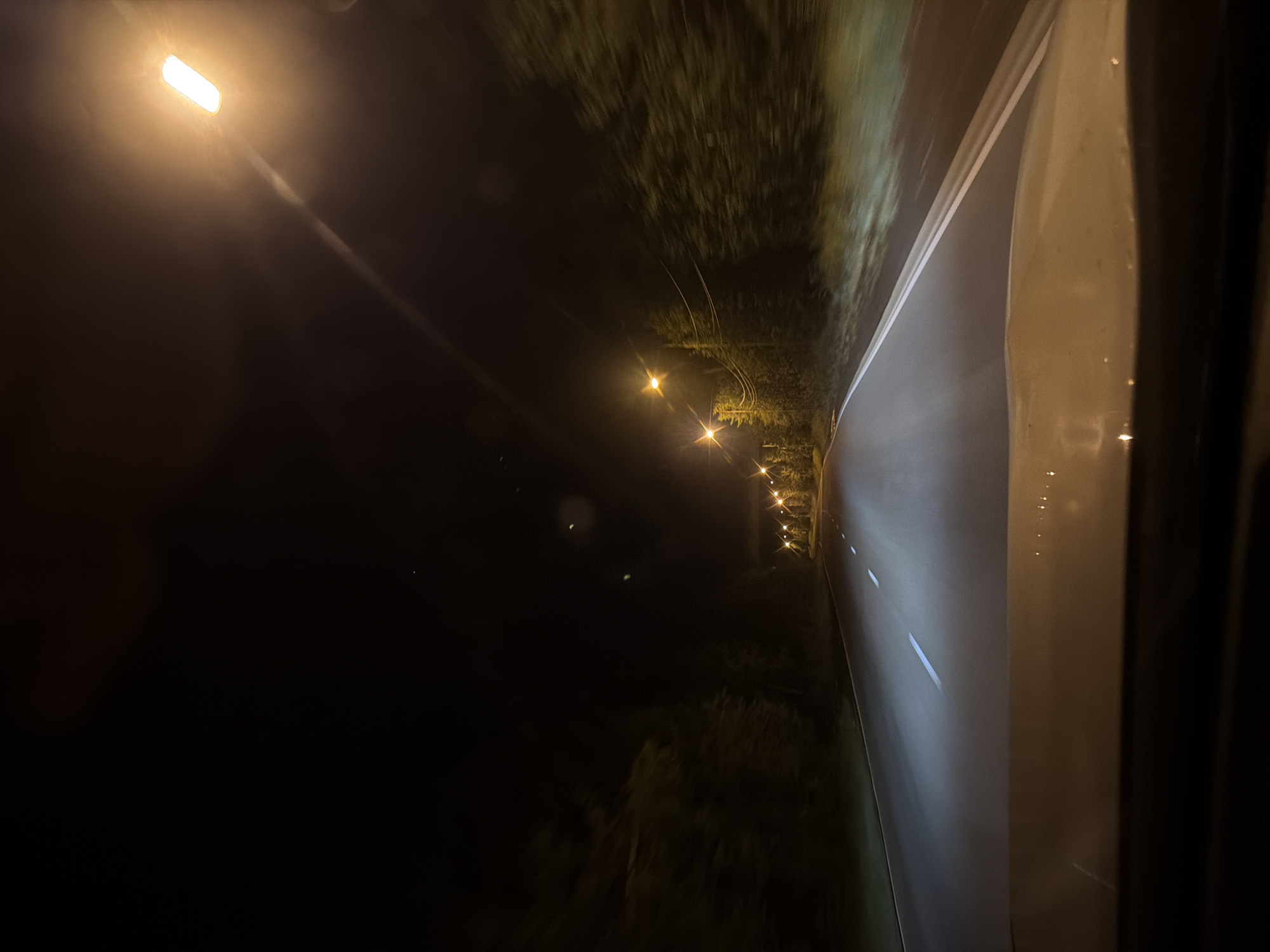 The image is a first-person perspective view from a vehicle driving at night on a paved road. The road is illuminated by streetlights and the vehicle's headlights. Trees line both sides of the road, blurred due to motion. The sky is dark, with no visible stars or moon. The dashboard of the vehicle is visible in the foreground, reflecting the light from the road and streetlights. The scene suggests a drive through a forested or rural area on the outskirts of a city. Based on the available information, this image could depict a road in Ust-Ilimsk, Russia, given the potential for nighttime driving in such locations.