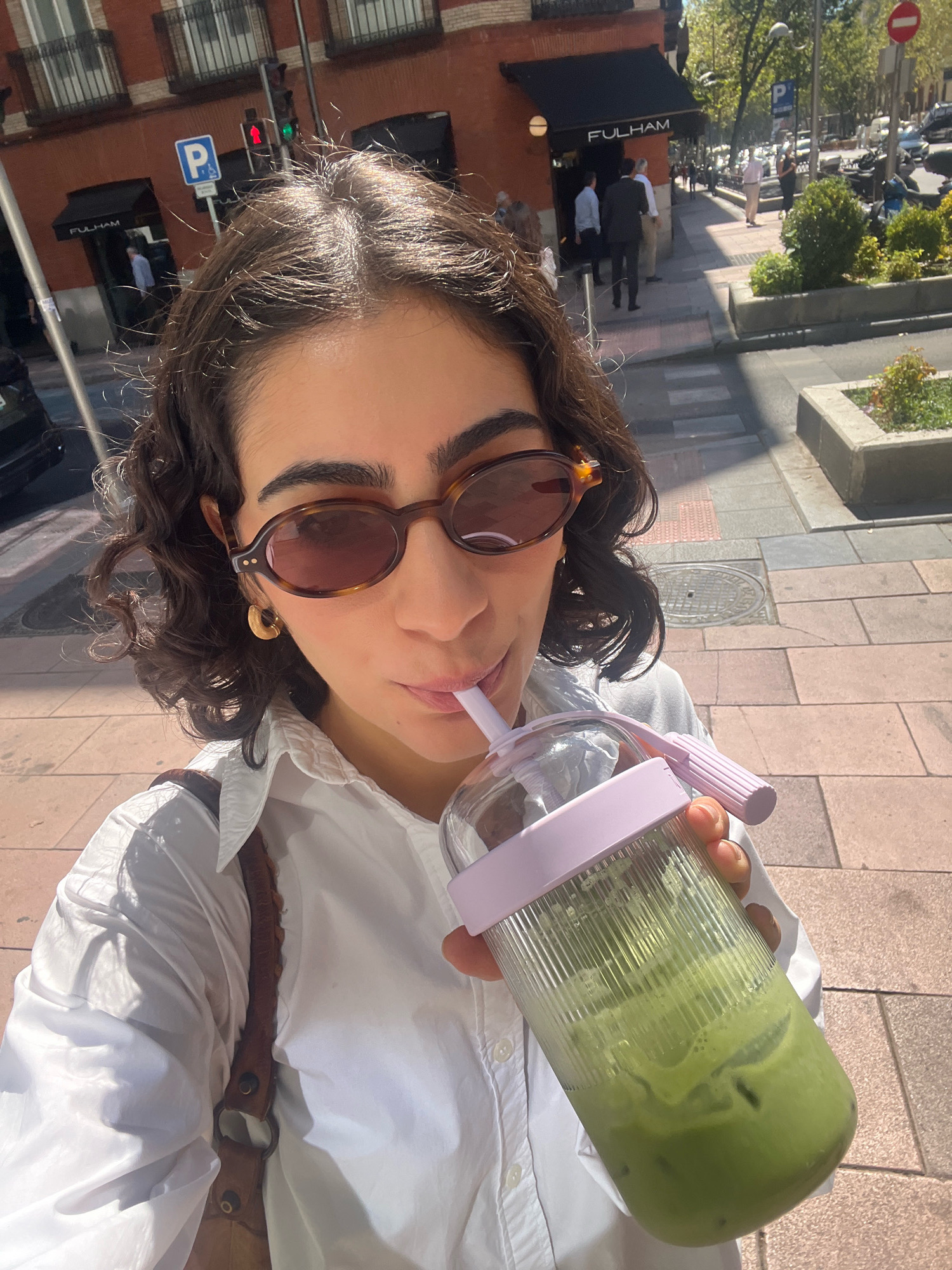 A person with curly brown hair and sunglasses is holding a clear plastic cup filled with a green beverage, possibly a matcha latte, and drinking it through a straw. The person is wearing a white collared shirt and a brown bag strap is visible over their shoulder. The background shows a street scene in Madrid, Spain, with a building labeled "FULHAM," a traffic light, a parking sign, and people walking on the sidewalk. The lighting suggests it is daytime.