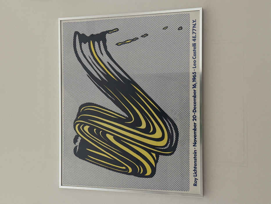 A framed print featuring a Pop Art style image hangs on a plain, light-colored wall. The artwork depicts a bold, stylized brushstroke in black and yellow, set against a grey background textured with a pattern of black Ben-Day dots. The brushstroke twists and turns from left to right, ending with black and yellow drips. Below the artwork, text in blue lettering reads: "Roy Lichtenstein • November 20 - December 16, 1965 • Leo Castelli 4E.77 N.Y." This identifies the artist, exhibition dates, and the Leo Castelli Gallery, located at 4 East 77th Street in New York, New York, United States of America. The frame is a thin, silver-colored metallic material. No people or other objects are visible in the scene.