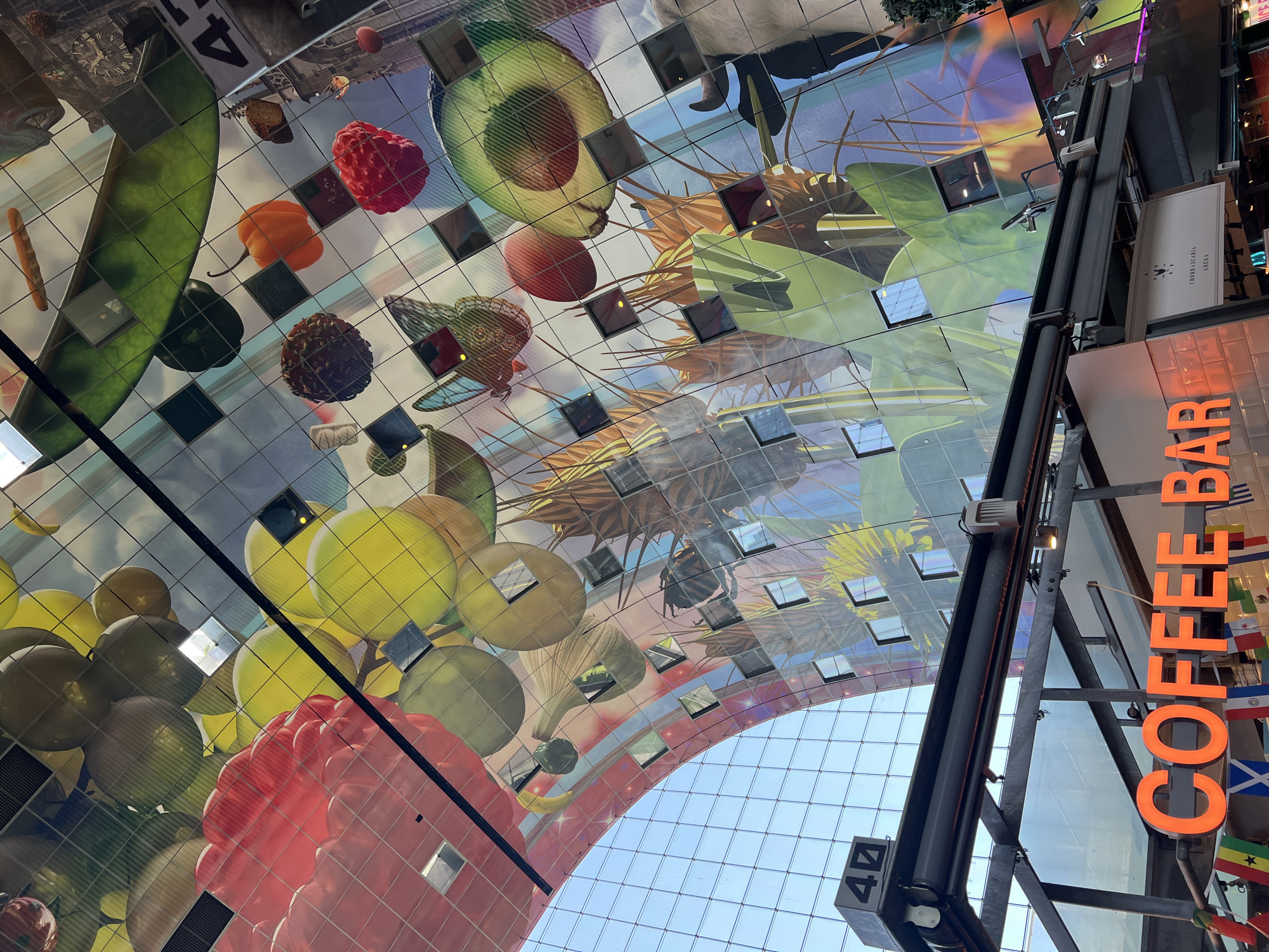 This is a low-angle, wide shot of the ceiling of a large, indoor market in Rotterdam, Netherlands, which is designed to resemble an open-air market with a curved, reflective ceiling covered in a vibrant mural of fruits, vegetables, and other food items. The mural is divided into square panels, some of which are reflective glass, creating a kaleidoscope effect.
The subjects of the mural include large illustrations of a green pea pod, raspberries, an avocado, oranges, grapes, and what appear to be pomegranates or tomatoes. There are also images of insects, possibly dragonflies or butterflies, and botanical elements like leaves and stalks.
The scene depicts the interior of a modern architectural space. The ceiling dominates the frame, and the camera is looking upwards towards it. The reflective nature of the ceiling panels allows for glimpses of the interior architecture and possibly some people or objects within the market, though they are distorted and fragmented.
Visible text includes the word "COFFEE" in large, orange, illuminated letters on a signpost on the right side of the image. Below the "COFFEE" sign, a smaller sign reads "CHURRASCARIA ADEGA". There is also a black box with the number "4" on it, visible in the upper left and lower left of the image.
The time of day appears to be daytime, indicated by the bright light illuminating the interior and the clear blue sky visible through the large, grid-like window structure of the market. The weather is not directly discernible but the interior setting suggests it is an indoor activity. The overall impression is one of a lively and visually engaging public space.