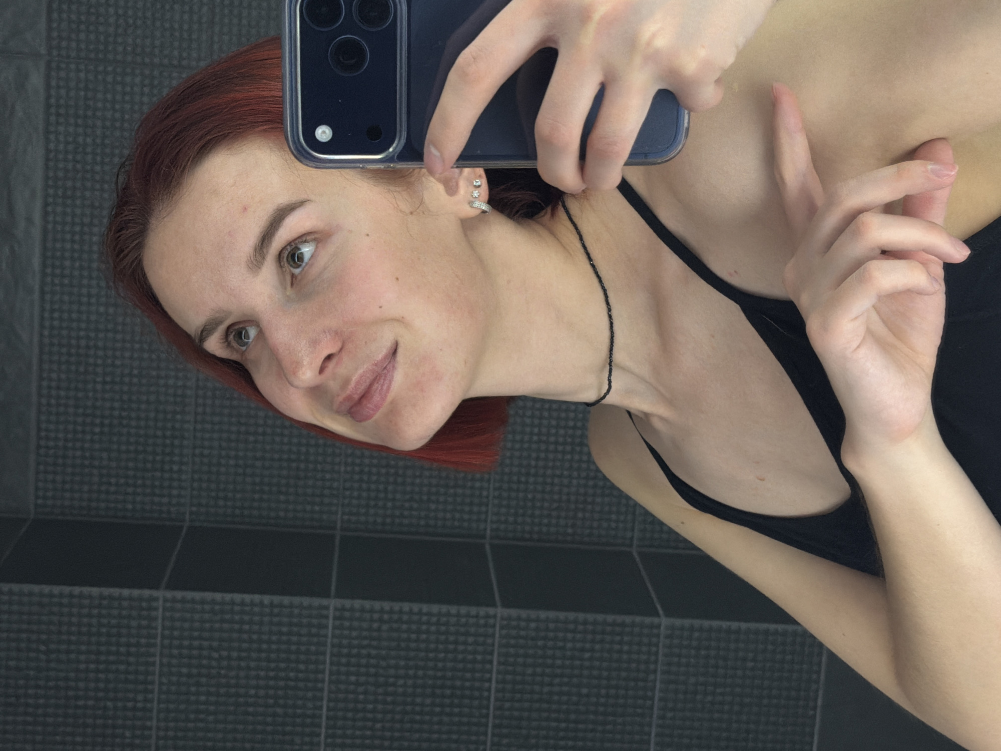 A selfie depicts a woman with reddish-brown hair, fair skin, and light eyes, holding a dark-colored smartphone with multiple camera lenses up to a mirror. She is wearing a black tank top and a delicate black beaded necklace. Three small earrings are visible in her left ear. The background shows a wall covered in small, dark gray square tiles. The photo appears to be taken in a bathroom. The location is Chaikovsky, Russia.