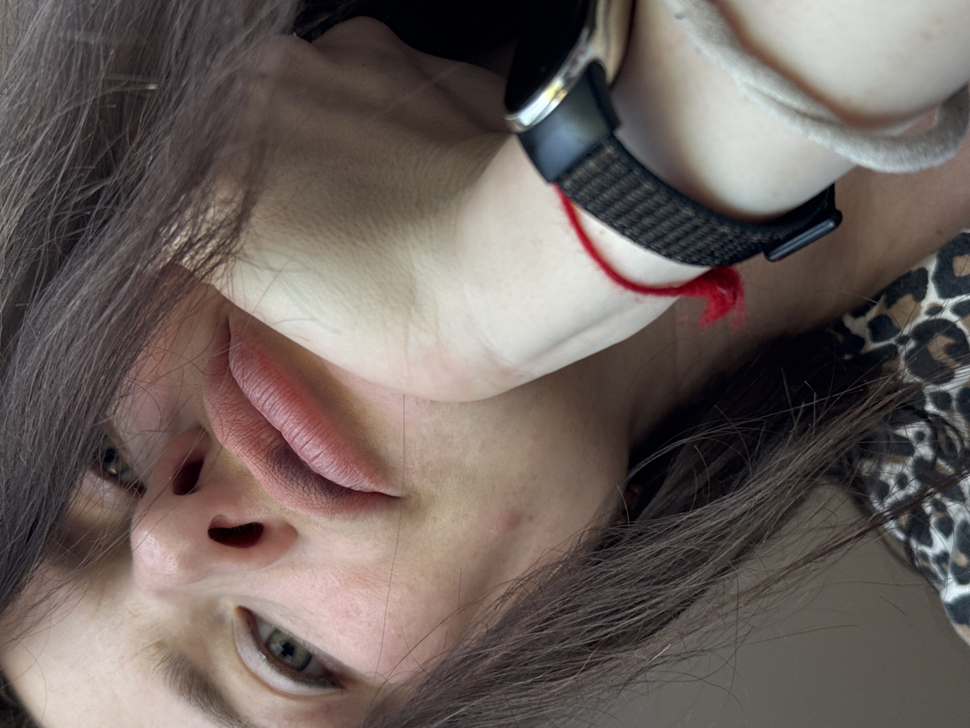 A close-up, slightly blurred, and rotated image captures a partial view of a person's face and neck. The individual has fair skin, brown hair, and light-colored eyes. A black smartwatch is visible on the wrist, accompanied by a light-colored rope bracelet and a red string. The person's lips are full and slightly parted. The background is out of focus. There is a glimpse of leopard print clothing on the side of the image. There is no information available in the image that places it in Rusalevka, Russia.
