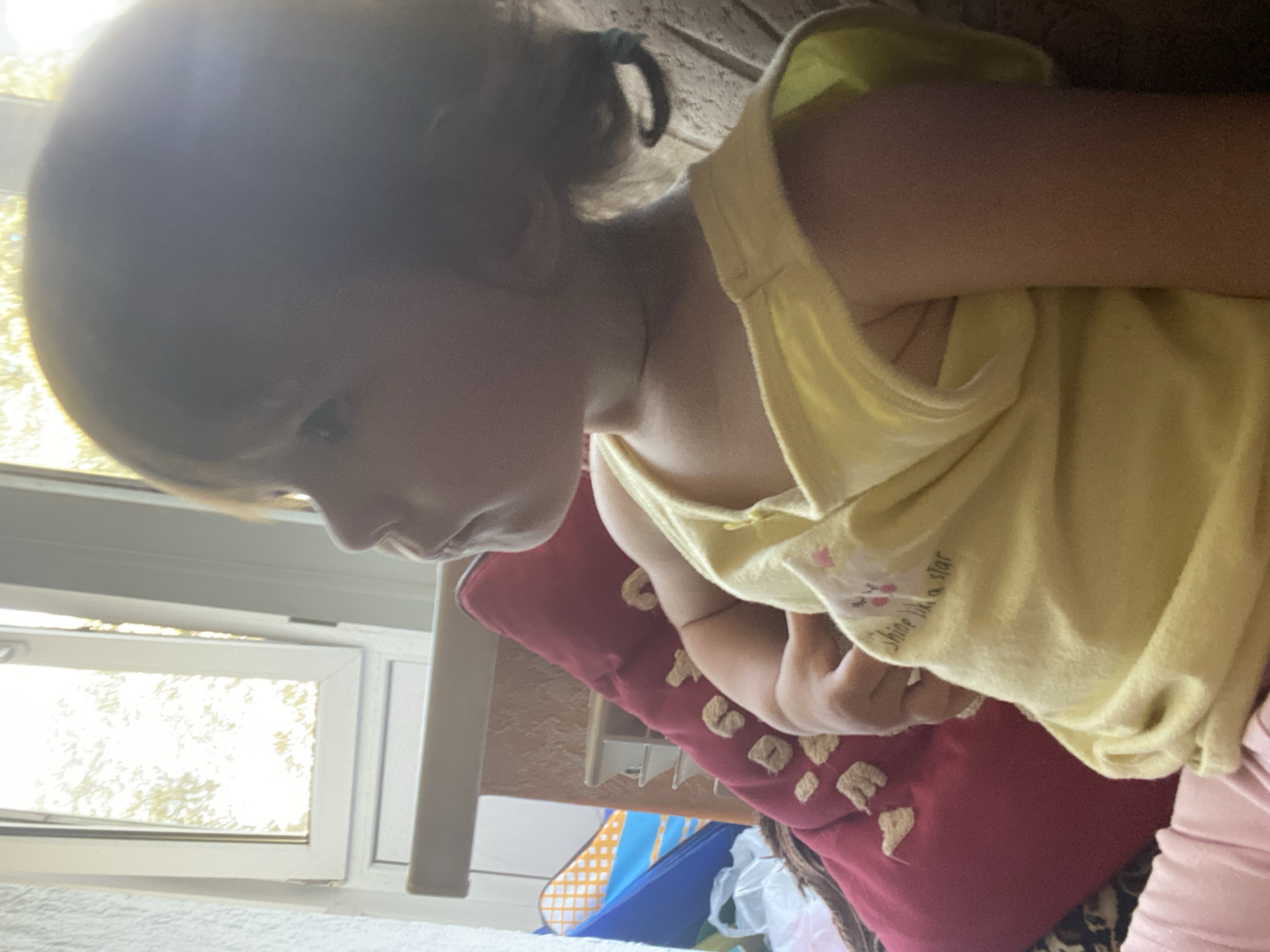 A close-up, profile shot captures a young girl with light brown hair, pulled back into a low ponytail with a blue tie, looking to her left with a pensive expression. She is wearing a bright yellow sleeveless shirt with a small graphic of a cat and the words "Shine like a star" printed on it. Her left arm is bent, and her hand rests on her lap. She is seated on a brown chair, with a textured pattern visible on the backrest. To her left, a large, plush red pillow with a pattern of beige paw prints is propped up. Behind the pillow, a blue and orange play tent is partially visible, along with some white plastic bags. In the background, a window with a textured glass pane lets in bright natural light, illuminating the scene. The overall environment appears to be indoors, possibly a living room or playroom, with a light-colored wall on the left.