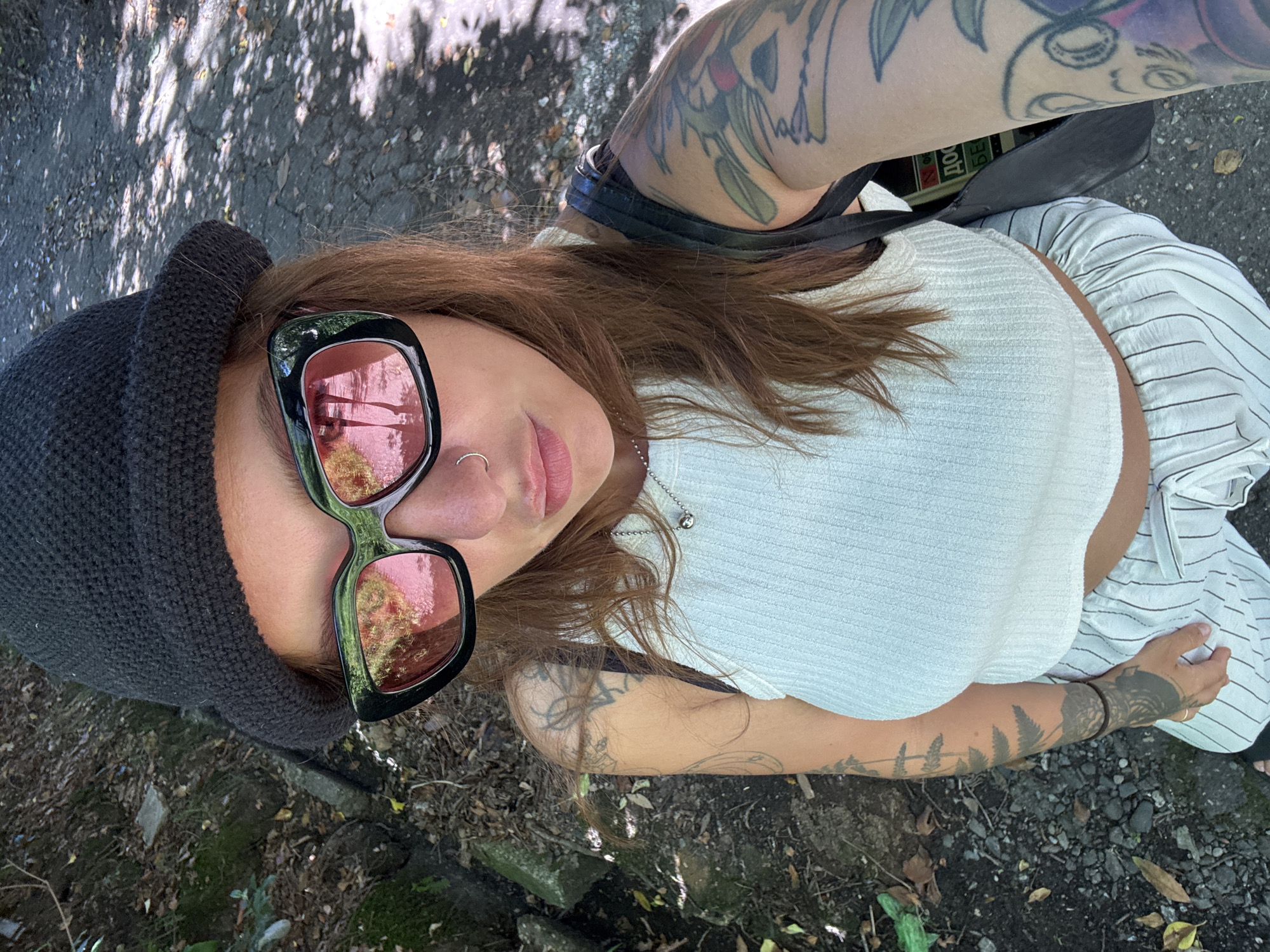 In an overhead, high-angle selfie, a woman stands in an outdoor, sun-dappled environment, possibly a park or walking trail in Sochi, Russia. She is wearing a black knitted beanie, large black-framed sunglasses with pink lenses that reflect trees, and a white ribbed crop top. Her lower half is obscured by a black strap of a bag and white striped pants. The ground is a mix of gravel and dirt, with scattered leaves and rocks, suggesting a natural, undeveloped area. The woman has multiple tattoos on her arms and a nose ring. The overall impression is of a casual, candid moment captured outdoors.