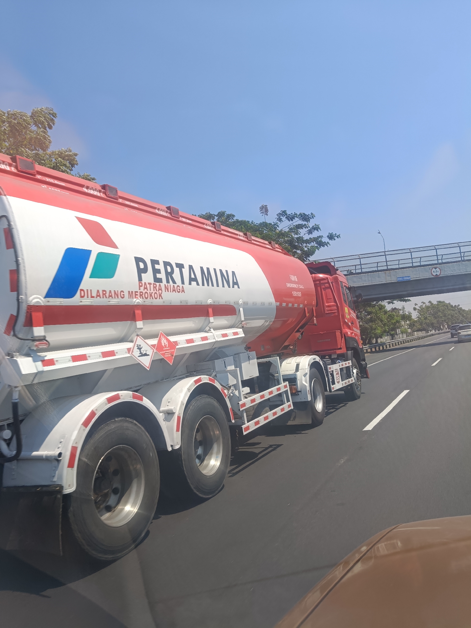In Sekarkurung, Indonesia, a red and white Pertamina tanker truck is driving on a highway under a clear blue sky. The truck has the Pertamina logo prominently displayed on its side, along with the text "PATRA NIAGA" and "DILARANG MEROKOK" (which translates to "No Smoking"). The truck also has hazard symbols indicating flammable materials. The highway has multiple lanes and is lined with trees and vegetation. An overhead pedestrian bridge is visible in the background, with a sign indicating a height restriction of 4.2 meters. Other vehicles are also present on the highway. The image appears to be taken from another vehicle moving alongside the tanker truck.