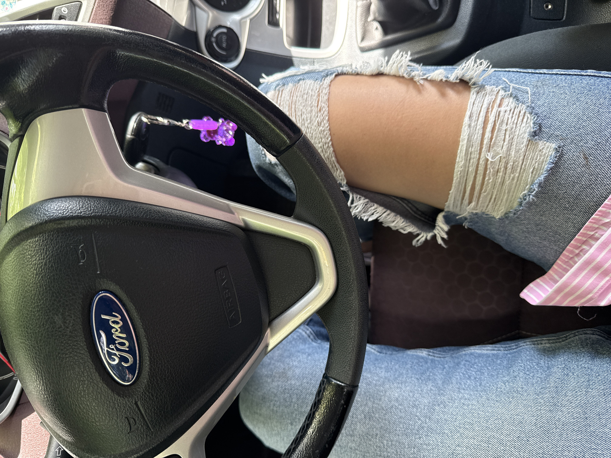 A first-person perspective shot shows a person sitting in the driver's seat of a Ford vehicle, with their legs visible wearing ripped denim jeans. A purple gummy bear-shaped keychain hangs from the ignition. The dashboard and steering wheel are visible, along with the center console. The overall setting appears to be inside a car.