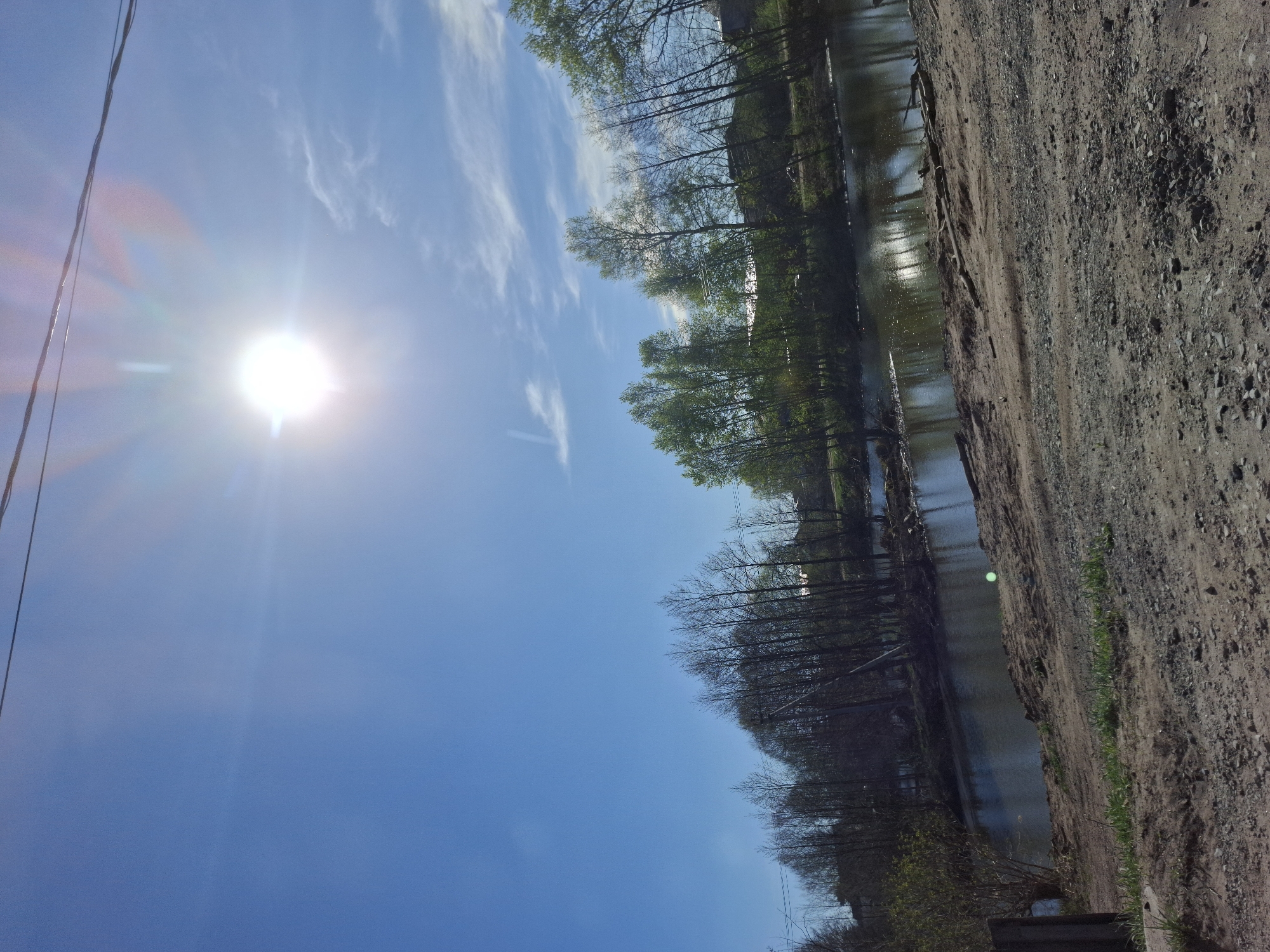 The image depicts a landscape in Chaikovsky, Russia. The sky is bright blue with a few wispy clouds and a blinding sun in the upper left. Powerlines are visible near the sun. A body of water, likely a river or lake, runs horizontally across the frame. Trees line the bank, some with leaves and some without. A dirt or gravel path runs along the right side of the water.