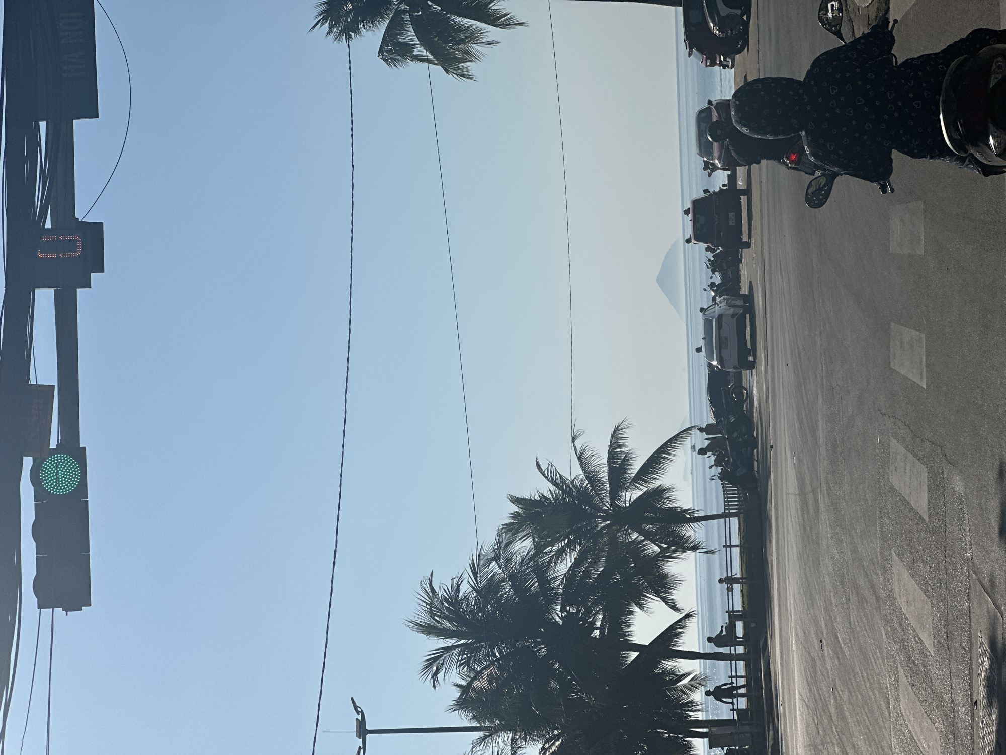 A street scene captured in Bac Nha Trang Ward, Vietnam. A traffic light shows a green signal. Palm trees line the street alongside the ocean. Vehicles are parked on the roadside. People are near the beach. In the distance, a mountain is visible.