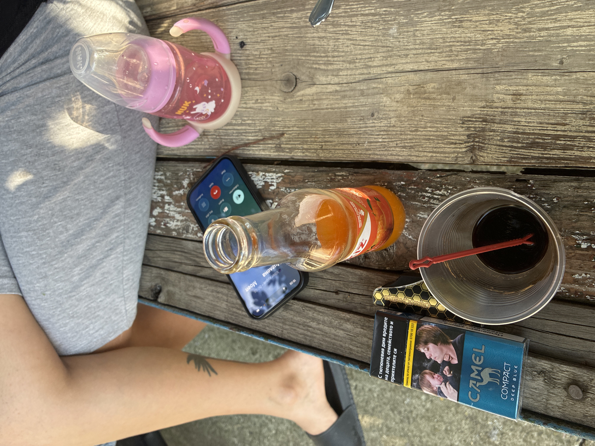 A high-angle outdoor shot captured in Radievo, Bulgaria, shows a weathered wooden plank table laden with various items, with the lower body of a person seated behind it. The individual wears grey shorts or sweatpants, with one leg crossed over the other. A dark tattoo, resembling a bird or feather, is visible on their left lower leg, which also wears a dark grey slip-on sandal.
On the table, from left to right, are:
* A blue "CAMEL COMPACT DEEP BLUE" cigarette pack, featuring a graphic health warning in Bulgarian depicting a crying child and an adult exhaling smoke.
* Next to the cigarette pack, a small object with a textured, golden-yellow and black honeycomb-like pattern, possibly a cigarette case or lighter.
* A clear plastic cup containing a dark brown liquid and a red plastic stir stick.
* A black smartphone, face-up, displaying a call screen with Cyrillic text and a "00:00 / 00:00" timestamp. The screen reflects ambient light.
* An amber glass bottle, partially filled with an orange liquid, resting beside the smartphone. Its label is partly visible, showing "UNITEME" and "200ml" text.
* A pink NUK baby bottle with handles, containing a pink liquid and decorated with white rabbit decals, positioned on the right side of the table.
* The tip of a silver spoon is visible on the far right edge of the table.
The wooden table surface exhibits prominent grain and wear, with chipped paint in areas. Sunlight casts shadows, notably on the person's clothing.