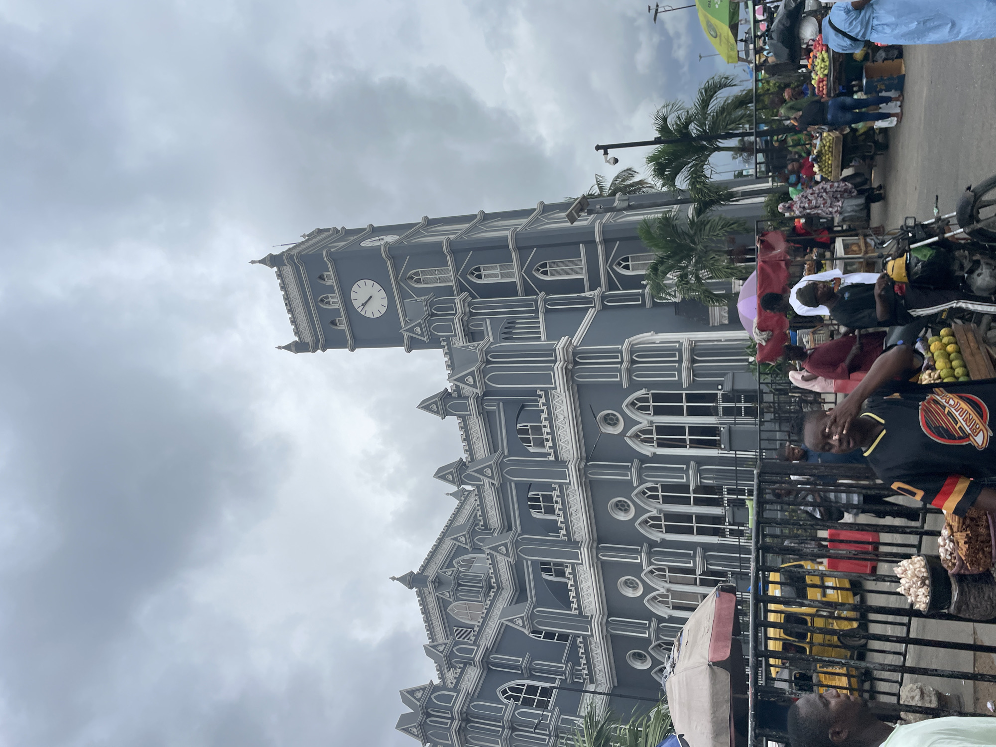 The image shows the Cathedral Church of Christ in Lagos, Nigeria, under a cloudy sky. The gray and white gothic-style architecture of the cathedral is prominent. In the foreground, a black metal fence separates the cathedral from a bustling market scene with people, motorcycles, fruit stands, and various goods for sale. Some people are visible behind the fence, including one wearing a black and gold basketball jersey. Overhead there are palm trees.