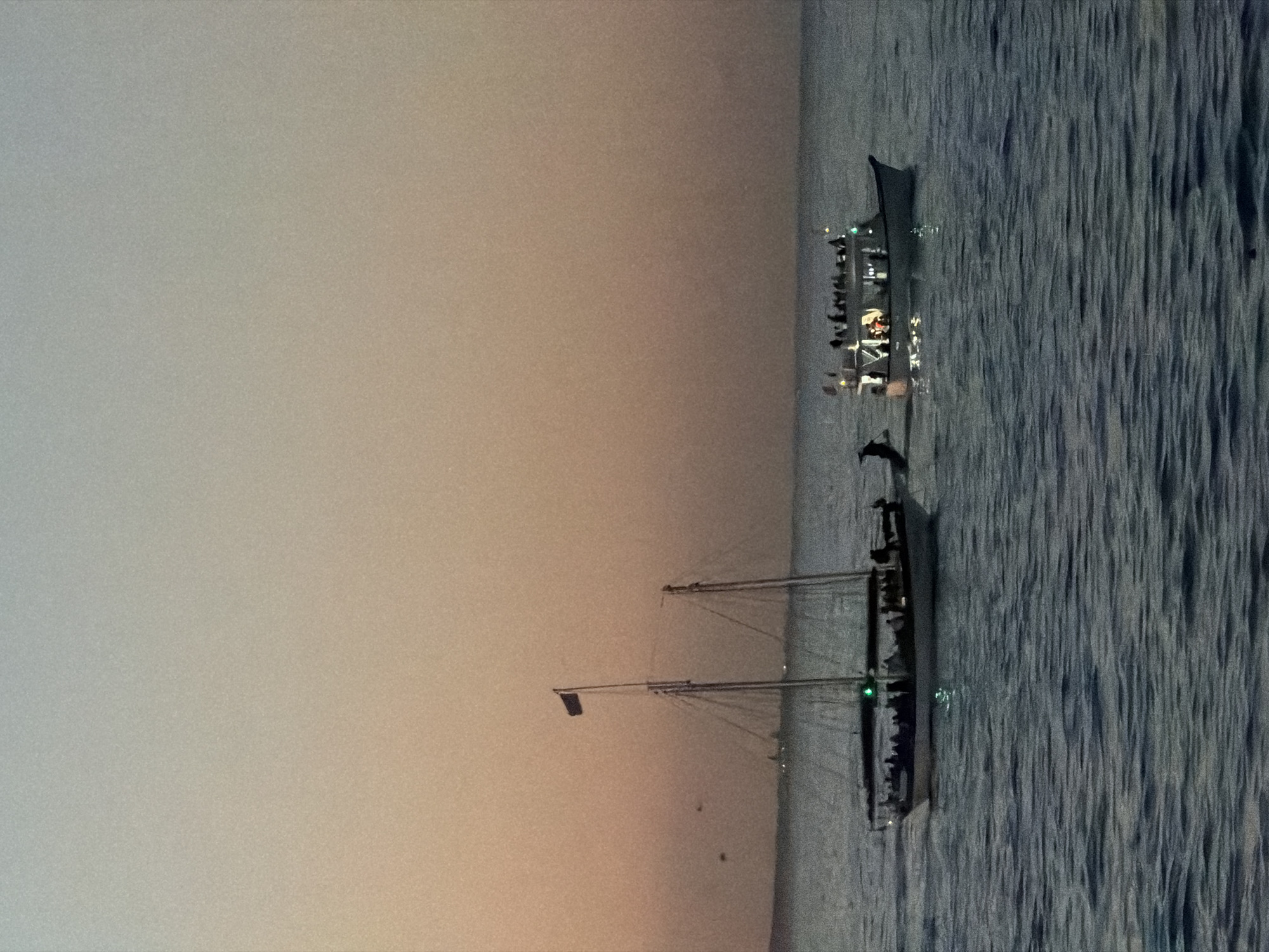 The image shows a nighttime scene of two boats on the water, likely at sea. In the background, there's another, smaller boat closer to the horizon with people on it. The water is visibly textured with wave patterns. The sky is a gradient from a pale orange/grey near the horizon to a darker shade above. Due to the lack of landmarks, confirming the location in Cape Town is impossible.