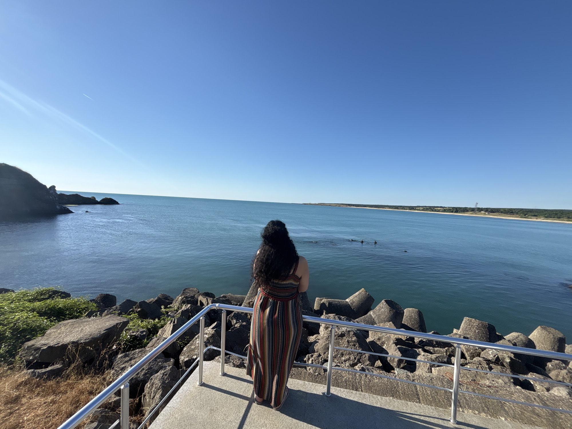 A woman with long, dark curly hair, wearing a long, multi-colored striped dress, stands with her back to the camera, looking out at the Black Sea from a concrete viewing platform in Rezovo, Bulgaria. The platform is bordered by a metal railing and a rocky shoreline with large, angular concrete blocks and natural rocks. The sea is calm and a vibrant turquoise blue. The sky is clear and a bright, cloudless blue, with a faint contrail from an airplane visible in the upper left quadrant. In the distance, a strip of land with trees and a structure resembling a tower can be seen. Several dark, rounded objects, possibly buoys or distant swimmers, are visible in the water. The sun is casting shadows on the platform.