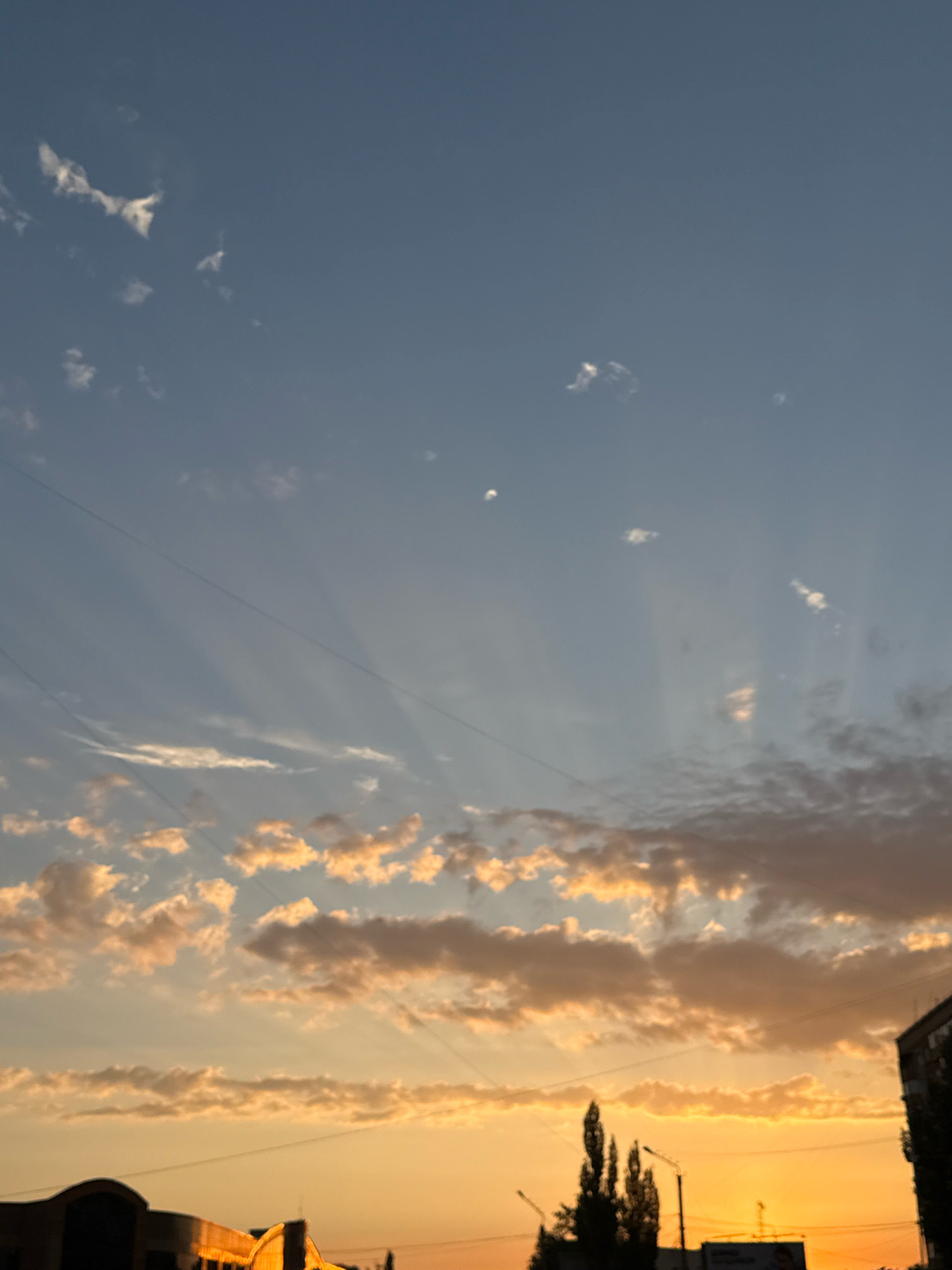 This image captures a sunset over the city of Pavlodar, Kazakhstan. The sky is a gradient of blue at the top, transitioning to a vibrant orange near the horizon. Wispy clouds, illuminated by the setting sun, are scattered across the sky. The sun's rays are visible as they pierce through the clouds, creating dramatic crepuscular rays.
In the foreground, the silhouettes of buildings and trees are visible against the bright sky. A power line is strung across the frame. The general ambiance is one of a tranquil evening in an urban setting. The image appears to be taken from a residential area, possibly an apartment building, given the perspective and the presence of power lines. The time of day is clearly sunset, with the warm hues dominating the scene. There are no discernible people or animals in the image. The overall scene suggests a peaceful conclusion to the day in Pavlodar.