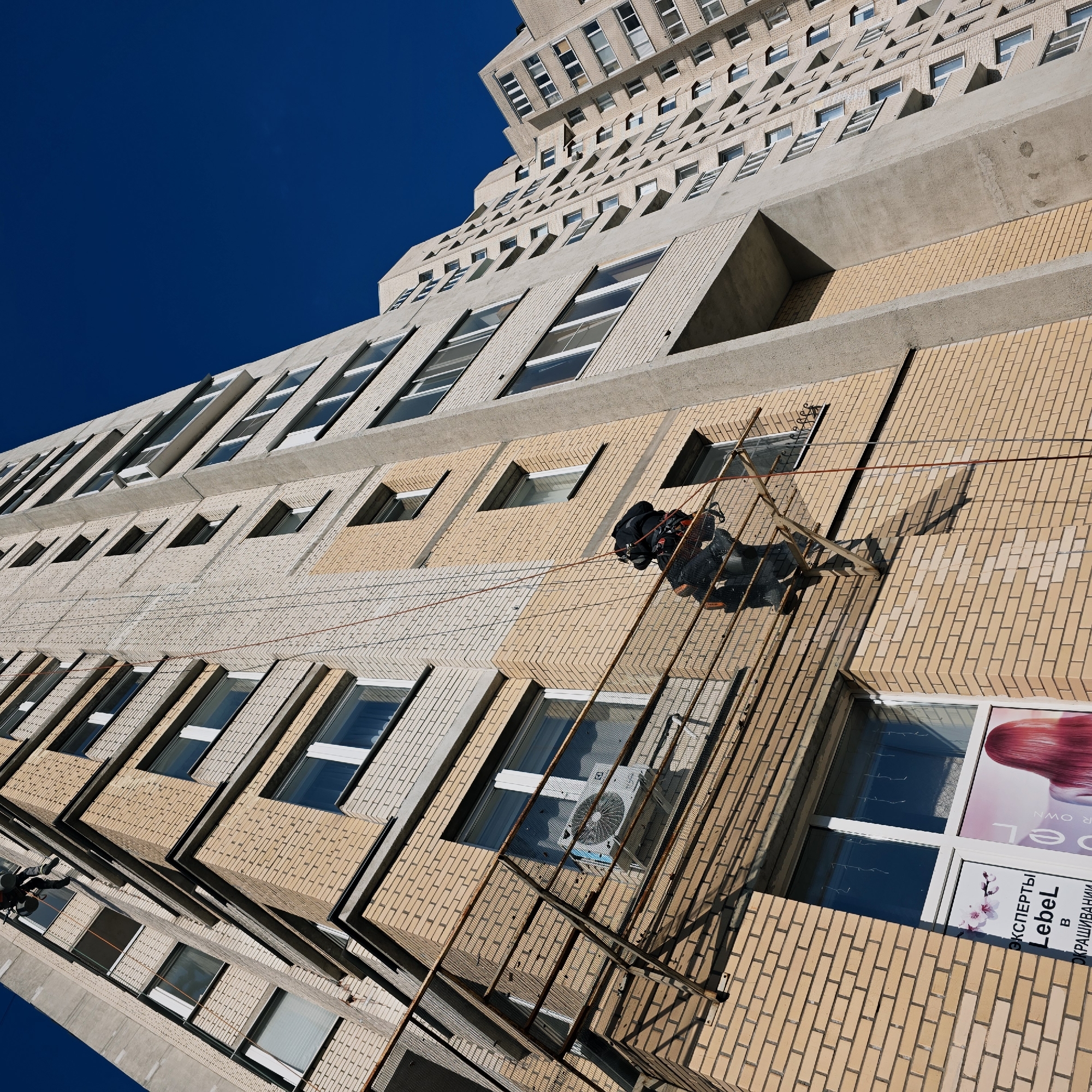 The image depicts an urban scene in Saint Petersburg, Russia, focused on a multi-story residential building under facade maintenance. The building features a mixed construction of light beige brickwork and grey concrete structural elements, with numerous windows. On the ground floor, a white-framed window displays a sign for "ЭКСПЕРТЫ LebeL в ОКРАШИВАНИИ" (LebeL Experts in Dyeing), indicating a hair salon, with graphics including cherry blossoms and a woman with red hair.
Two individuals identified as industrial climbers are visible on the building's facade. In the foreground, one climber, dressed in dark workwear with orange highlights on the knees/shins and wearing a harness, is suspended by red ropes. This climber is positioned approximately on the third floor, standing on or leaning against a rusty metal grid structure that protrudes from the building, potentially supplemented by a wooden platform. The climber appears to be actively working on the facade. Below a window at this level, a white exterior air conditioning unit is mounted. Higher up and to the left, a second climber, also in dark clothing and attached to ropes, is partially visible, indicating ongoing work on a higher section of the facade.
The building extends upwards, revealing many floors with various windows and small balconies or decorative protrusions. Another, more distant multi-story building with similar architecture is visible in the background to the right. The sky is clear and deep blue, suggesting a sunny day, with pronounced shadows cast on the building's facade.
