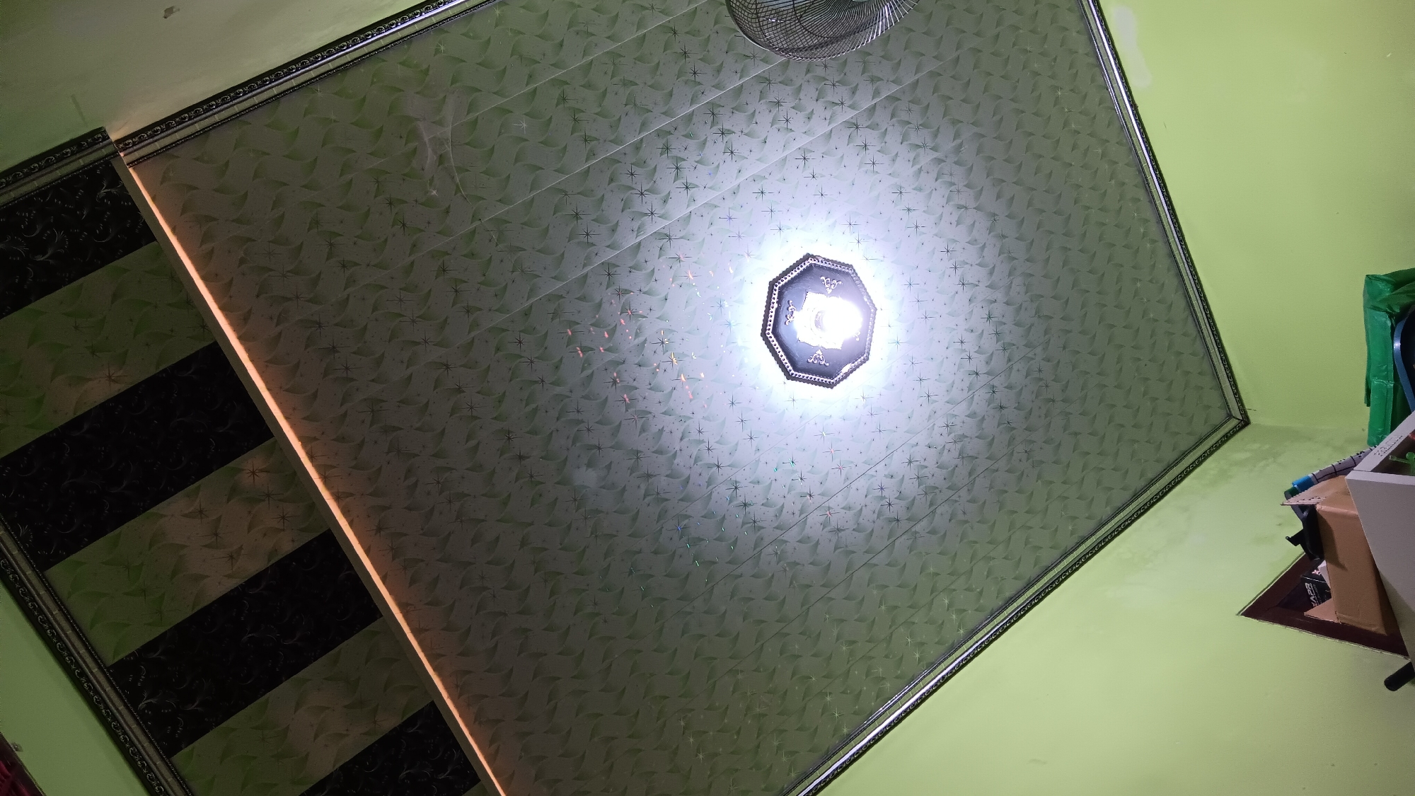 The image captures a low-angle view of a room's interior, focusing on the ceiling and upper walls. The ceiling is covered with patterned tiles, predominantly light green with a repeating geometric design that resembles abstract flowers or stars. A central ceiling light fixture, octagonal in shape with a dark frame, is illuminated brightly, casting a white glow that diffuses across the ceiling. To the left, a section of the ceiling features black and green stripes with a similar repeating pattern, separated by a thin, illuminated strip of light, possibly an LED strip. The upper walls are painted a light, vibrant green. In the lower portion of the image, the corner of the room is visible, with a glimpse of shelves and some stacked boxes, suggesting a storage area. A part of a metallic fan grille is visible on the far right. The scene appears to be an indoor residential or commercial space. The image does not provide any specific information that can be used to determine its exact location in Sonobekel, Indonesia.