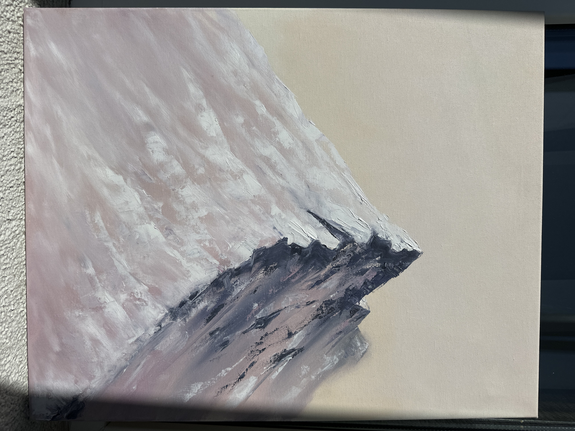 The image shows an abstract painting on canvas depicting a mountain. The mountain occupies the left side of the canvas, with a predominantly white and pinkish-gray color palette. The right side of the canvas is filled with a plain beige background. The painting exhibits textured brushstrokes, particularly in the mountain area, suggesting a thick application of paint. The scene is indoors. The location is unknown.