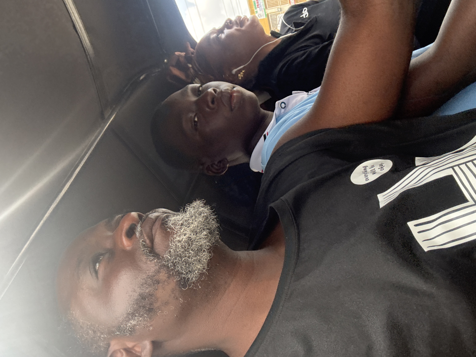 In Ado, Nigeria, three people are seen inside a motorized tricycle. The person in the foreground, a man, is looking up and to the right, his face partially obscured by a graying beard. He is wearing a black t-shirt with a graphic design on it. To his right, a young person in a light blue polo shirt with white and red trim looks directly at the camera. Behind them, a woman with earrings and a black top is visible in profile, looking to the right. The interior of the tricycle's roof is visible at the top of the frame. The lighting suggests it is daytime.
