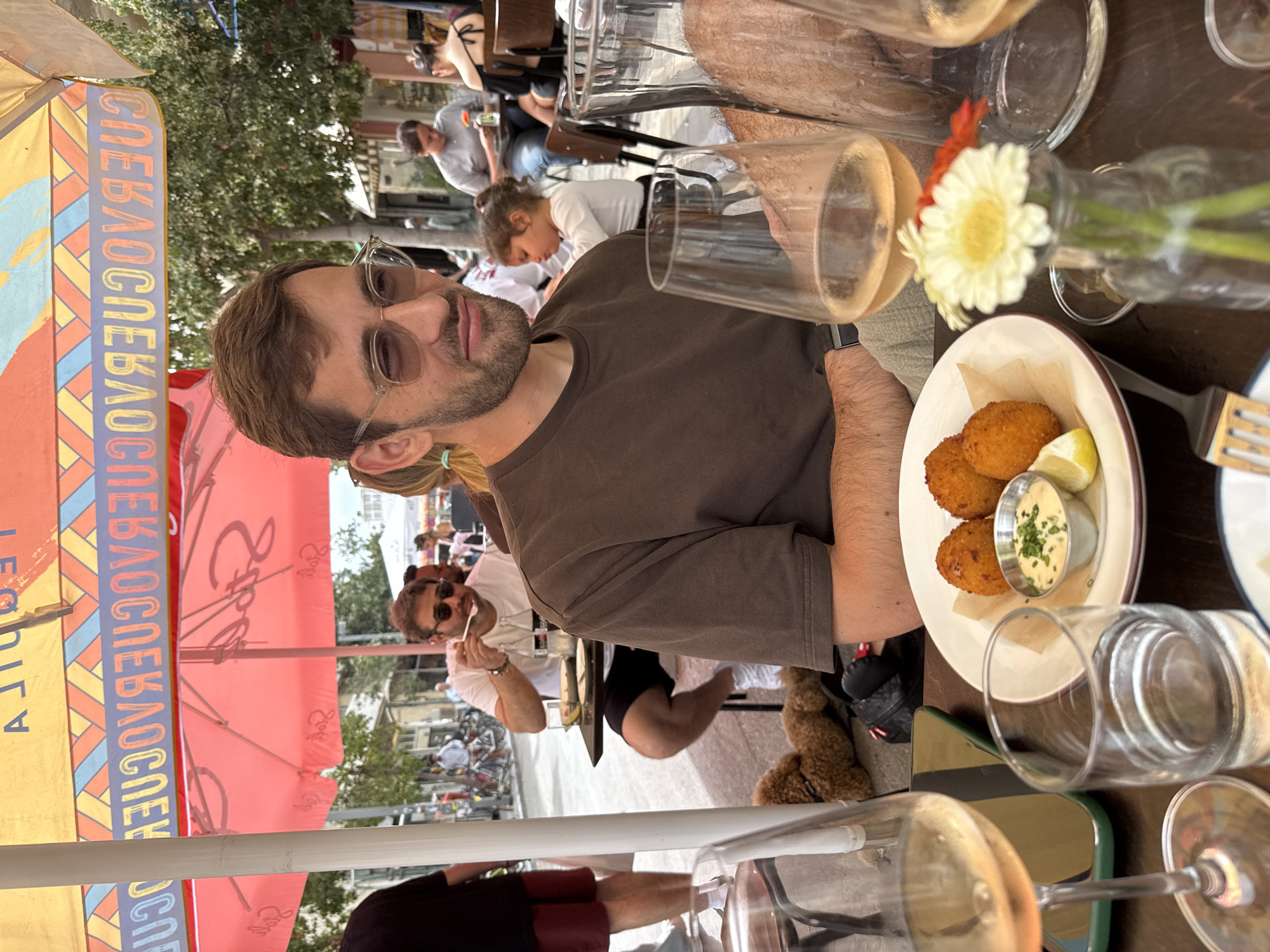 The image depicts a man with a beard and glasses seated at a table outdoors, possibly in a restaurant or cafe in Tel Aviv, Israel. The setting appears to be a bustling urban environment, as evidenced by the presence of other people, umbrellas, and street furniture. The man is wearing a brown t-shirt. On the table, there is a plate with a fried dish and a dipping sauce, a vase with flowers, and several glasses. The scene is brightly lit, suggesting it is daytime.