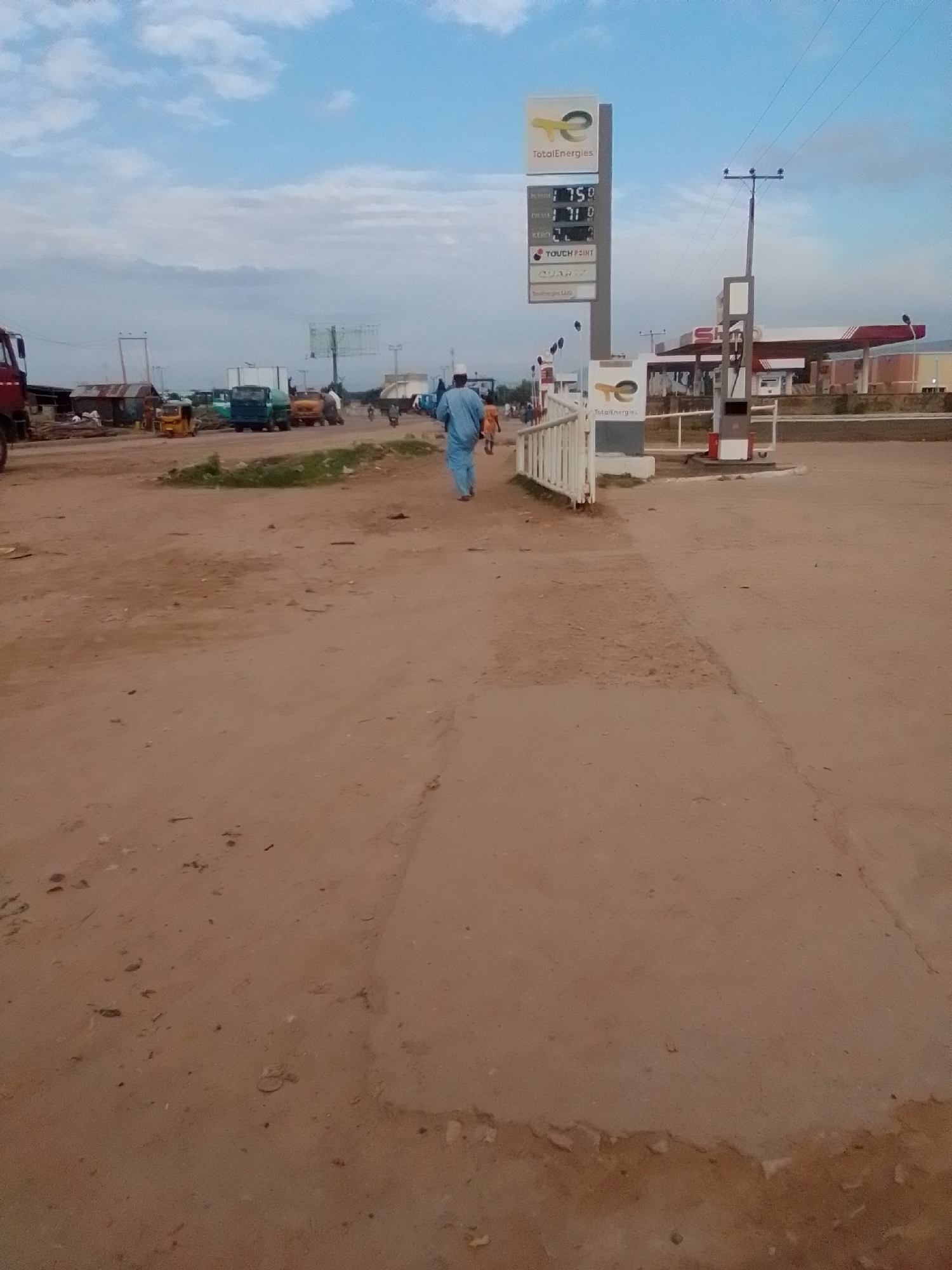 The image depicts a street scene in Gombe, Nigeria, near a TotalEnergies fuel station. The sky is partly cloudy, with a mix of blue and white clouds. A prominent TotalEnergies sign stands tall on the left side of the frame, displaying fuel prices: 750 for petrol, 710 for diesel, and 2 for kerosene. Beneath the prices, "TOUCH POINT" and "QUARTZ" are visible. Further down the road, the fuel station canopy is visible, with pumps and signage. Several vehicles are present, including a red truck on the far left, various trucks and a yellow tricycle in the middle ground, and a motorcycle in the distance. People are seen walking and standing around, including a man in a blue robe and white head covering walking towards the foreground. The ground is unpaved, with a dusty, reddish-brown surface. A white fence or barrier is located in front of the fuel station entrance. The overall impression is that of a busy, everyday scene at a commercial area in Gombe.