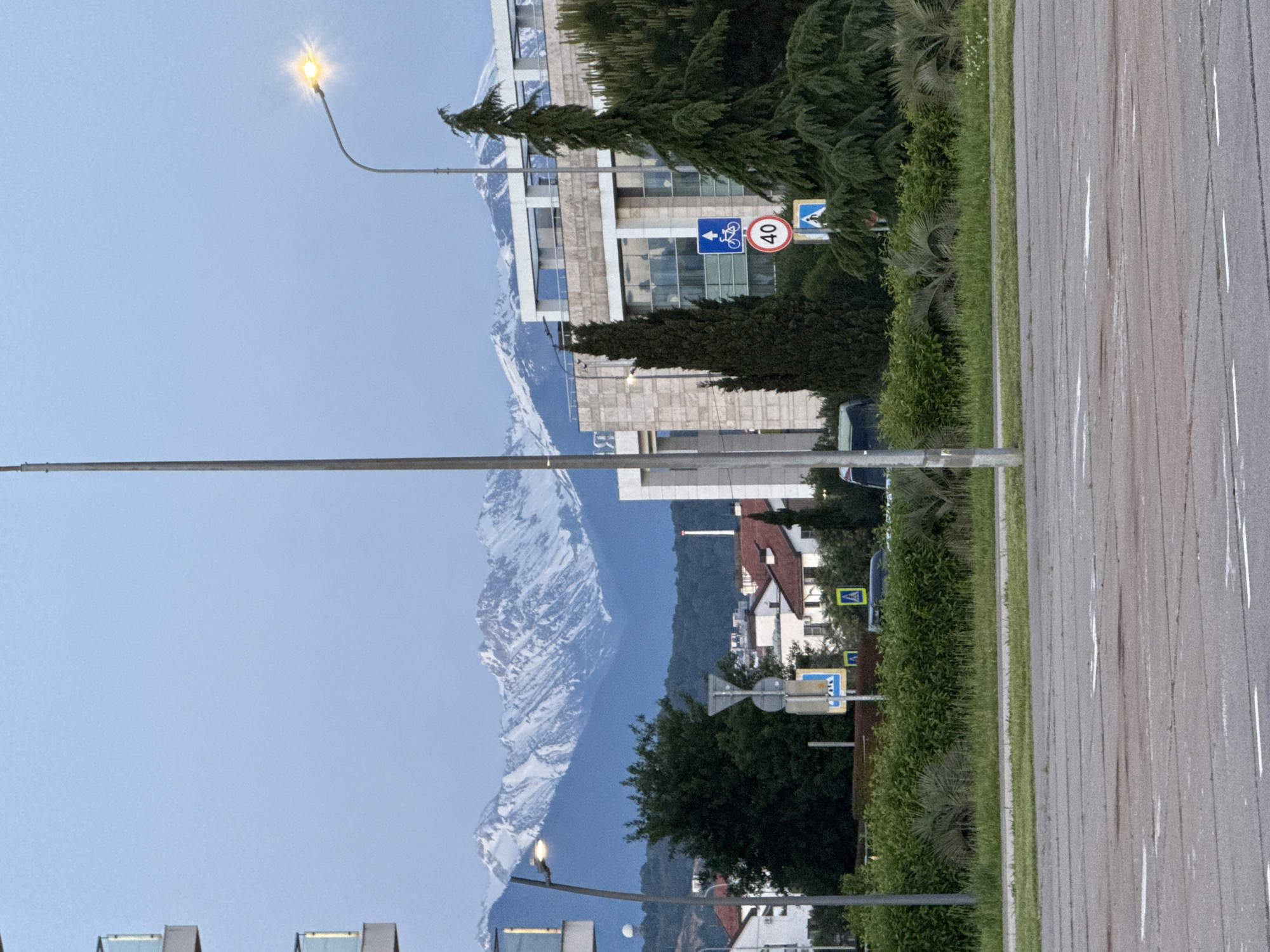 This image depicts a vibrant urban landscape in Sirius, Russia, situated against a dramatic backdrop of towering, snow-capped mountains. The foreground features a multi-lane road on the right, distinguished by white lane markings. Alongside the road, a green strip of grass, dense bushes, and some palm-like plants create a lush barrier. Several utility poles and streetlights, one of which is visibly glowing, are also present.
In the midground, a mix of architectural styles is visible, including modern, multi-story buildings with large windows and what appear to be residential structures with reddish-brown tiled roofs. Tall, dark evergreen trees, possibly cypresses, are interspersed among the buildings and abundant foliage, contributing to the green aesthetic.
Key traffic signs are noticeable: a blue rectangular sign with a white bicycle symbol and a left-pointing arrow, a circular red and white speed limit sign displaying "40", and yellow diamond-shaped and blue square signs indicating pedestrian crossings.
The distant background is dominated by rugged mountains, their upper slopes covered in glistening snow under a clear, light blue sky. The stark contrast between the warm, green, and developed foreground and the cold, majestic snow peaks is striking. The bright, clear sky and general lighting suggest it is daytime, with the glowing streetlight adding a touch of ambiance. The scene captures a bustling yet scenic environment, characteristic of a location where urban life meets impressive natural beauty.