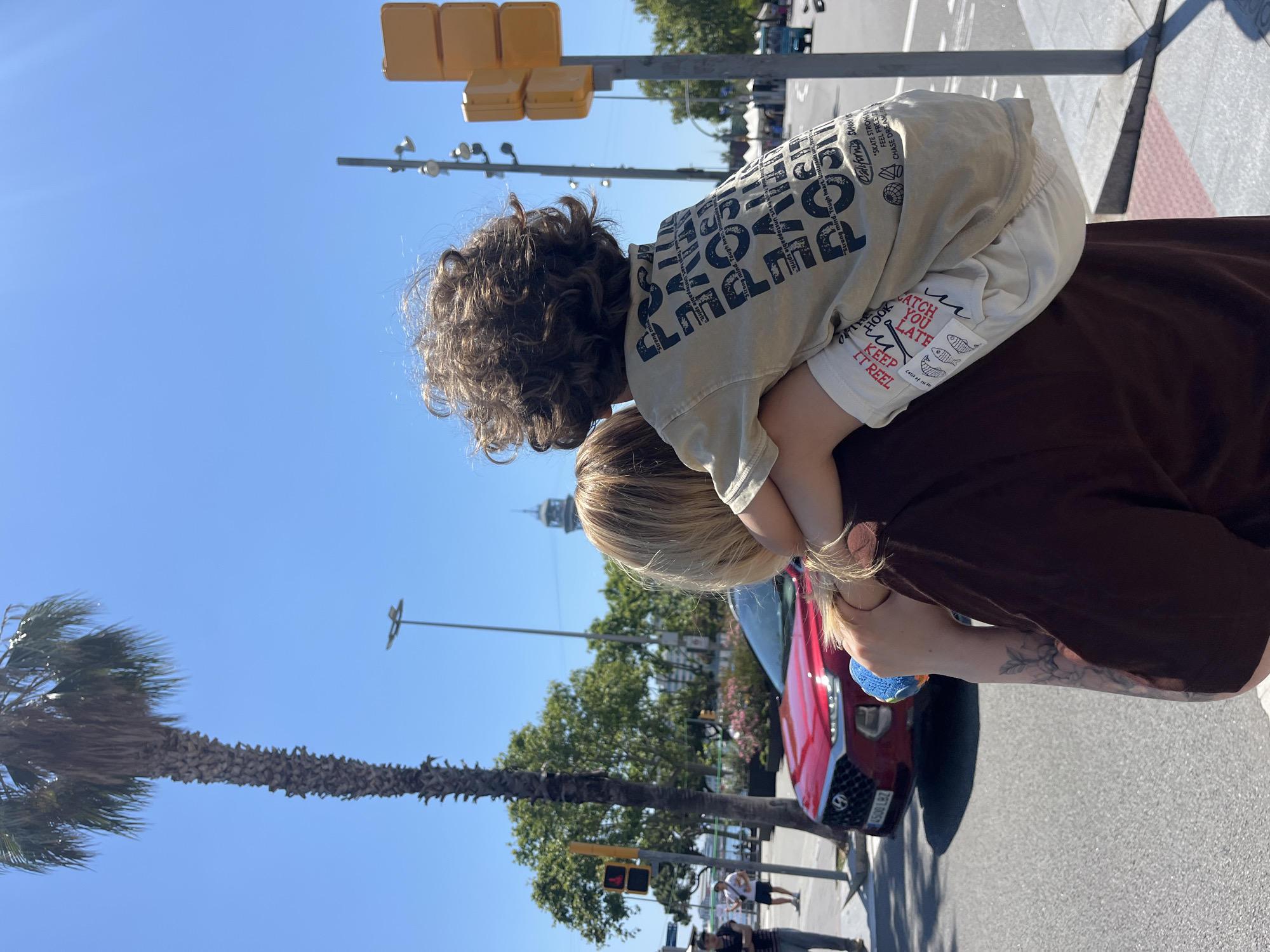 A vertical outdoor medium shot shows a child on the shoulders of an adult, likely the parent, on the street. They are likely in Barcelona, Spain, because a tower and a palm tree typical of Barcelona, Spain are visible in the background. The child has curly dark brown hair, and wears a tan t-shirt with words on it. The adult has blond hair and wears a brown t-shirt. Both are facing forward in the frame, though their bodies are angled slightly. Behind them is a red SUV, and a pedestrian walk signal light, palm trees, and a few other people walking. The sky is blue.