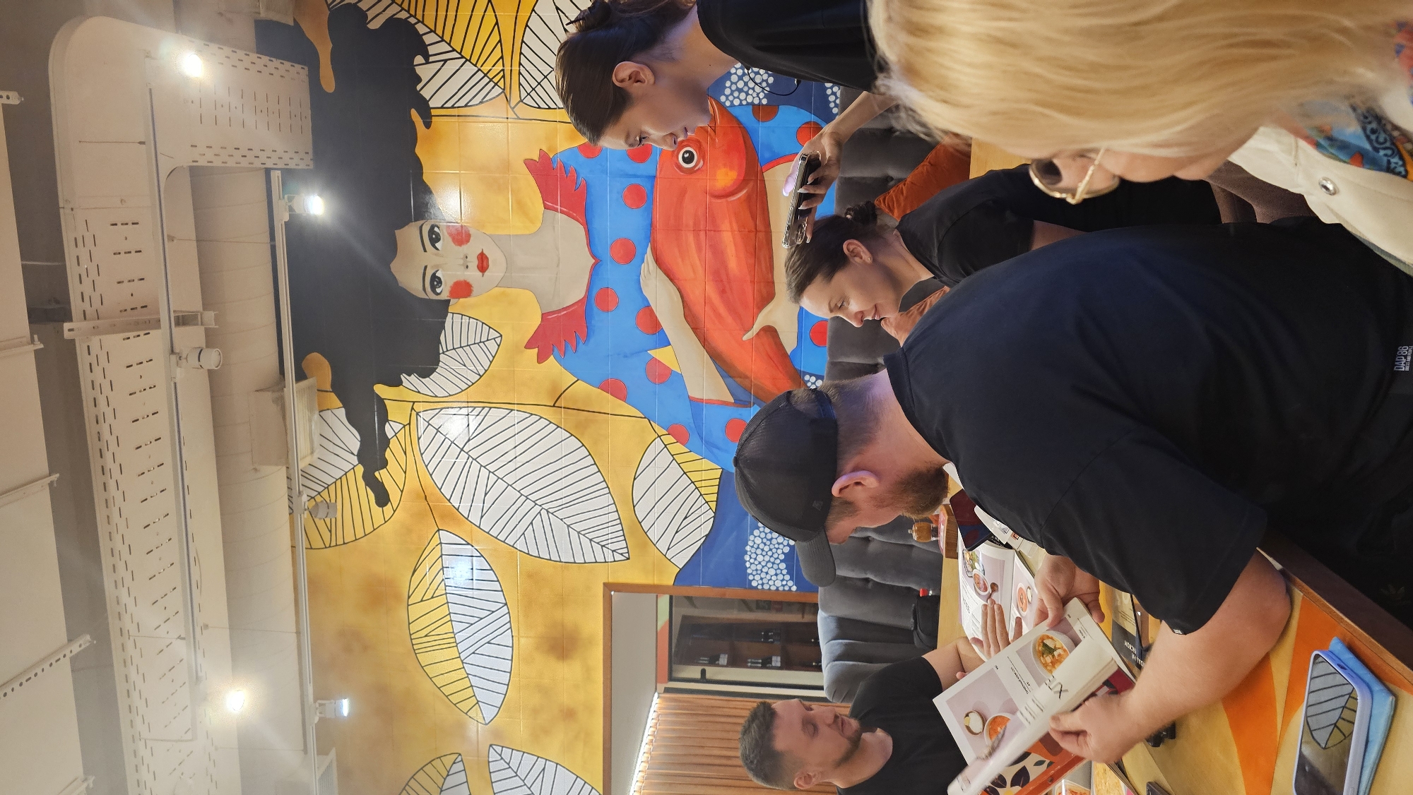 A group of people are gathered at a table, intently examining a menu, seemingly deciding on their meal choices. They are in a restaurant. The background features a mural with an artistic style, including a portrait of a person with striking red lips and a large orange fish. The mural suggests an international or Asian fusion style. The lighting is bright, which helps with the visual appeal of the mural. The specific location of the city of Sochi, Russia is not discernible based on the visual elements provided.