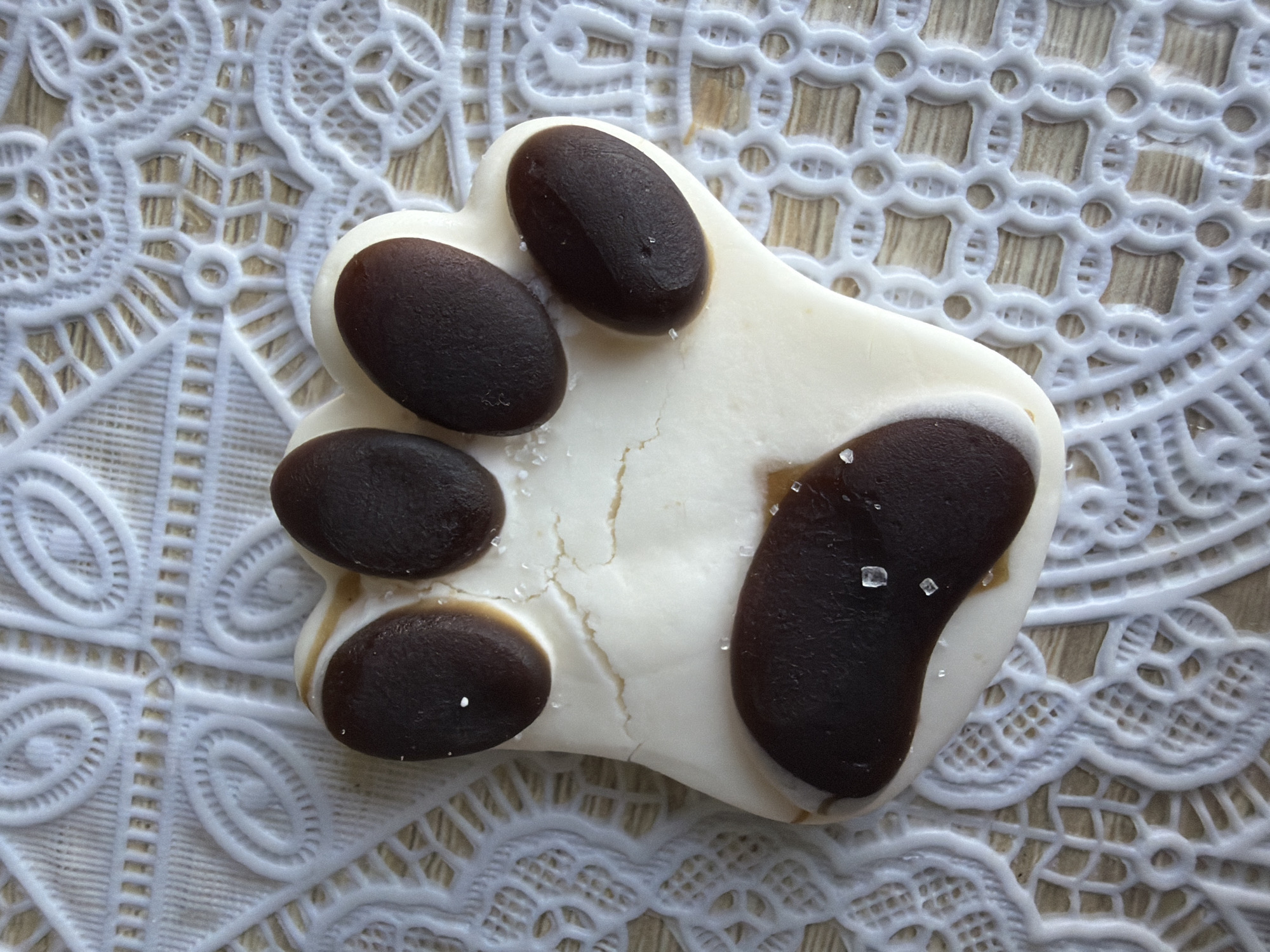 The image shows a close-up, overhead view of a decorative white lace placemat. On the placemat lies a paw-shaped cookie made of white frosting with dark brown candy "pads". The cookie has five "pads" arranged in a typical paw formation: four smaller ones at the top, representing toes, and one larger one at the bottom, representing the palm. Some of the brown pads appear slightly irregular in shape, and there are a few small white sugar crystals sprinkled on the cookie and the placemat. The background of the placemat appears to be a wooden surface. No people or actions are visible in the image. The location in the city of Strășeni, Moldova is not discernible from the visual information provided.