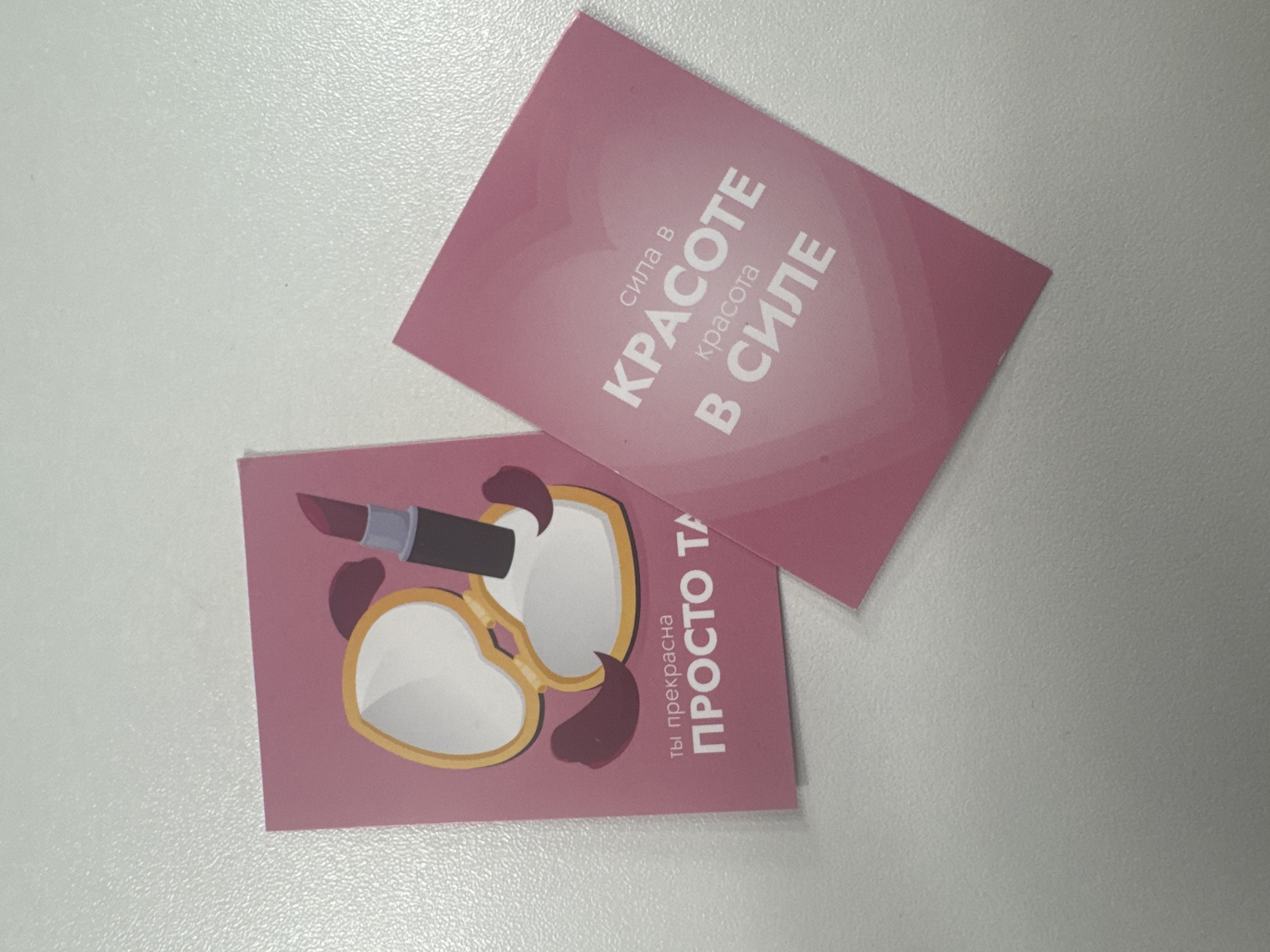 The image shows two promotional cards on a white surface. The top card is primarily pink and contains Russian text which translates to "Strength in BEAUTY, Beauty in STRENGTH." The bottom card, also pink, features a lipstick and a stylized pair of heart-shaped glasses. The text on the lower card translates to "You are beautiful. Simply." The setting is unknown and there is nothing in the image to suggest the location of Saint Petersburg, Russia.