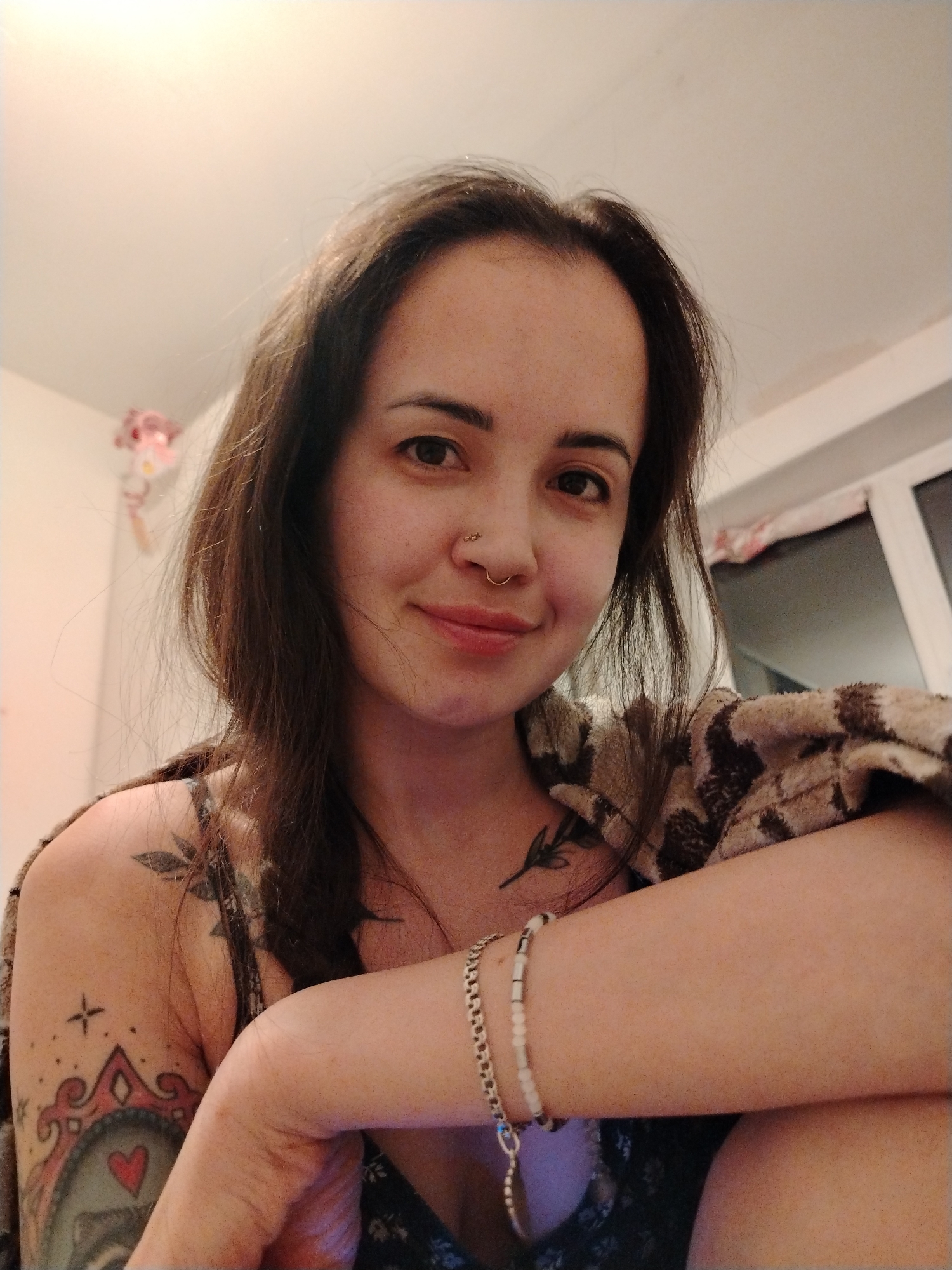 A woman with long, dark hair and a nose ring is looking at the camera with a slight smile. She is wearing a patterned top and has a blanket or shawl draped over her shoulders. Her left arm is visible, adorned with a bracelet and several tattoos. The background is somewhat blurred, but a window and a wall are discernible. It is not possible to confirm the location is Noviy, Russia.