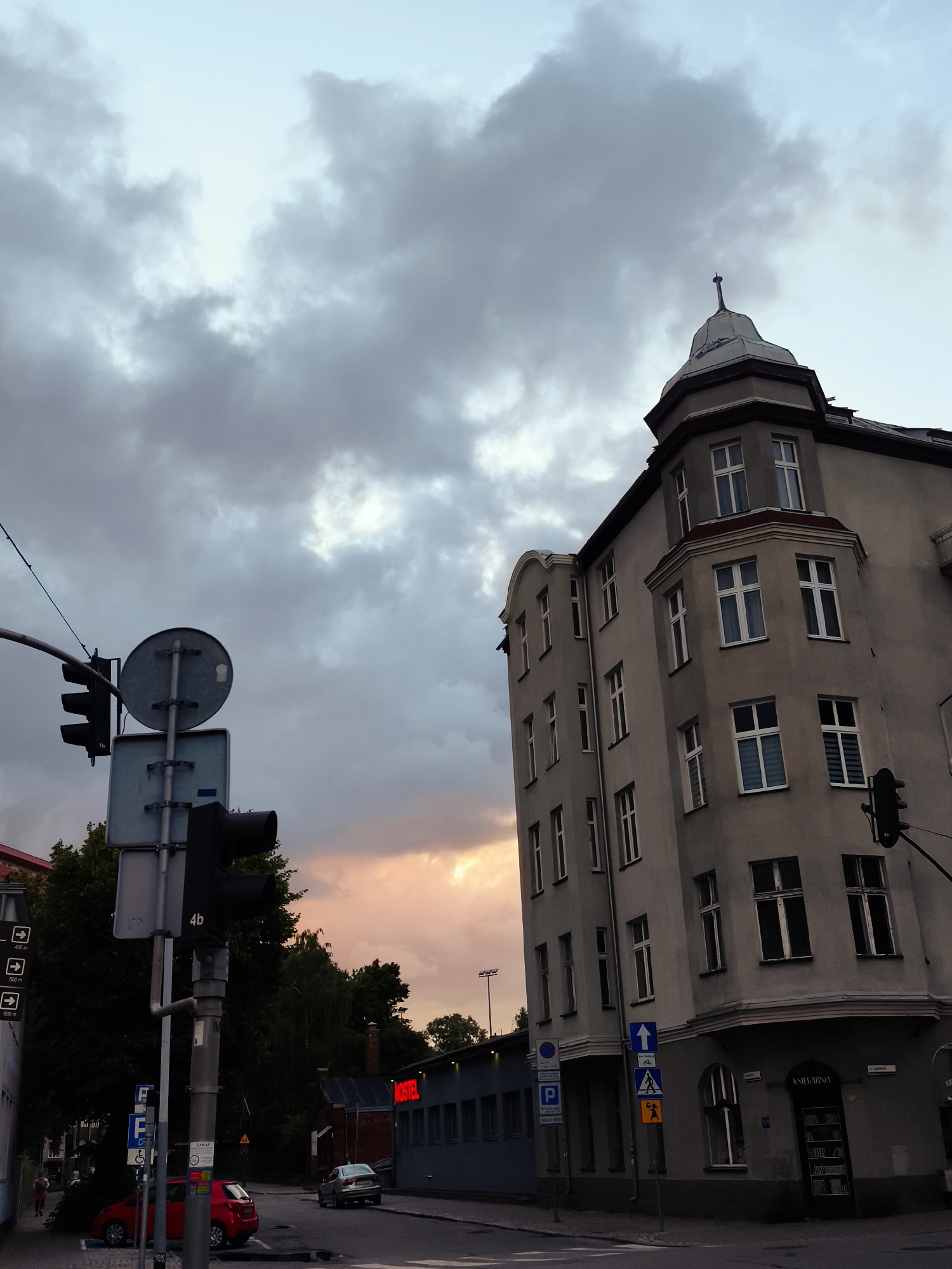 The image captures an outdoor urban scene, likely during twilight, with a focus on a street corner. The sky is filled with dramatic, dark clouds that are partially illuminated by a peachy sunset glow breaking through. Dominating the right side of the frame is a tall, multi-story building with a distinctive rounded corner and a small turret-like structure at its apex. The building's facade is a neutral gray, punctuated by numerous rectangular windows, some with white frames and others appearing to have curtains.
In the foreground, a traffic light stands tall, its signal heads turned away from the viewer. Several street signs are mounted on the same pole, including directional signs and a sign with the number "4b". To the left of the traffic light, a traffic signal with three lights is visible.
A red car is parked on the left side of the street, and further down, a silver car is driving away. A blue building with a red sign that reads "HOSTEL" is visible in the background, to the right of the trees. The street is paved, and a sidewalk is present. The overall impression is that of a somewhat moody evening in a European city.
The location is identified as Gdańsk, Poland, specifically at the intersection of Ulica Mikołaja Reja and Ulica Wita Stwosza. The building on the corner is the historic building at Ulica Mikołaja Reja 25, often referred to as the "Dom pod Kolumną" (House under the Column). The bookstore visible on the ground floor of this building is "Księgarnia Elżbietańska".