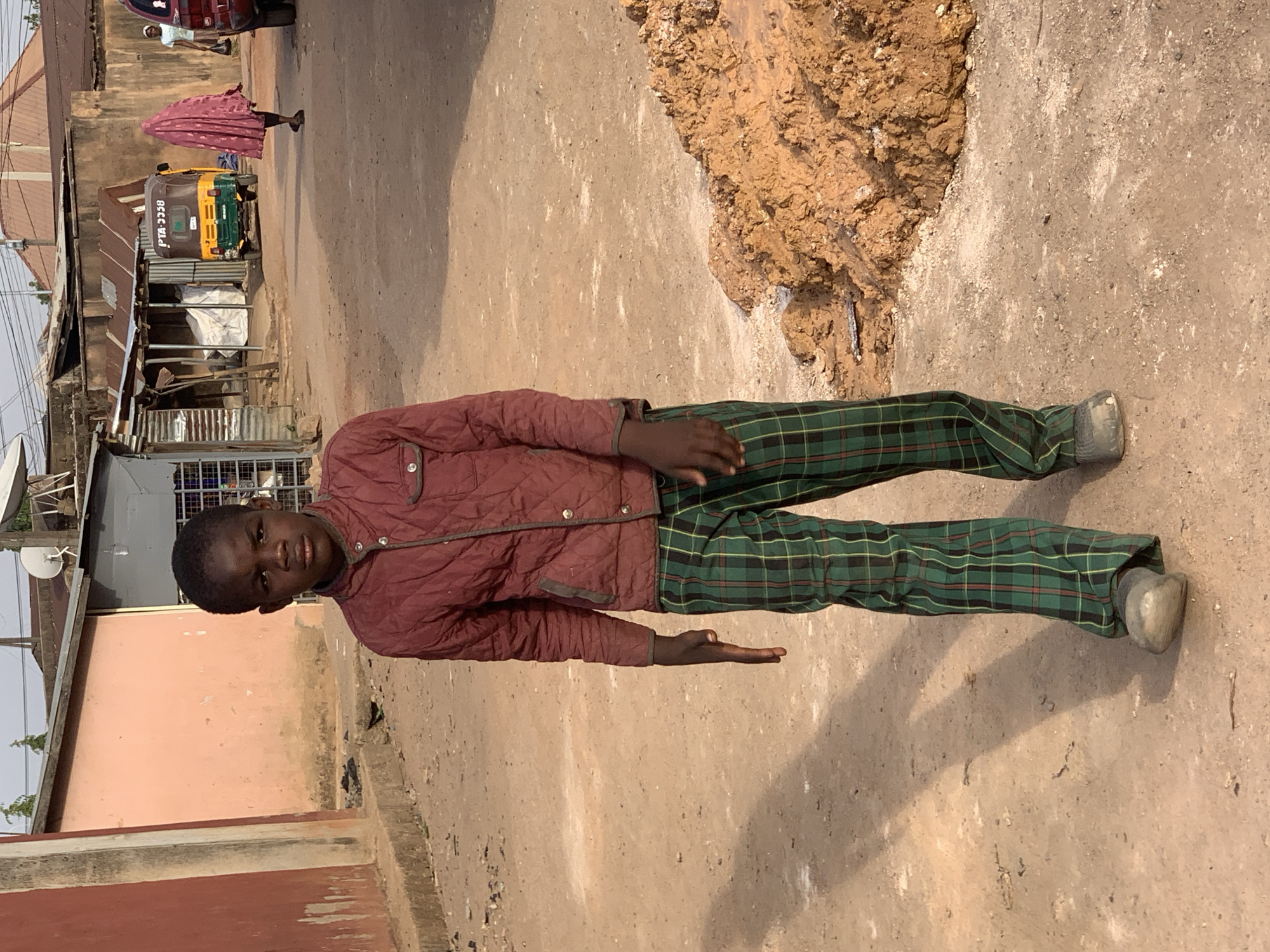 A boy in a red jacket and green plaid pants stands on a dirt road in Jos, Nigeria. He is facing the camera and his right arm is extended. To his right is a pile of dirt. Behind him, a woman in a pink head covering walks past a yellow and green tricycle. A building with a satellite dish is visible in the background.