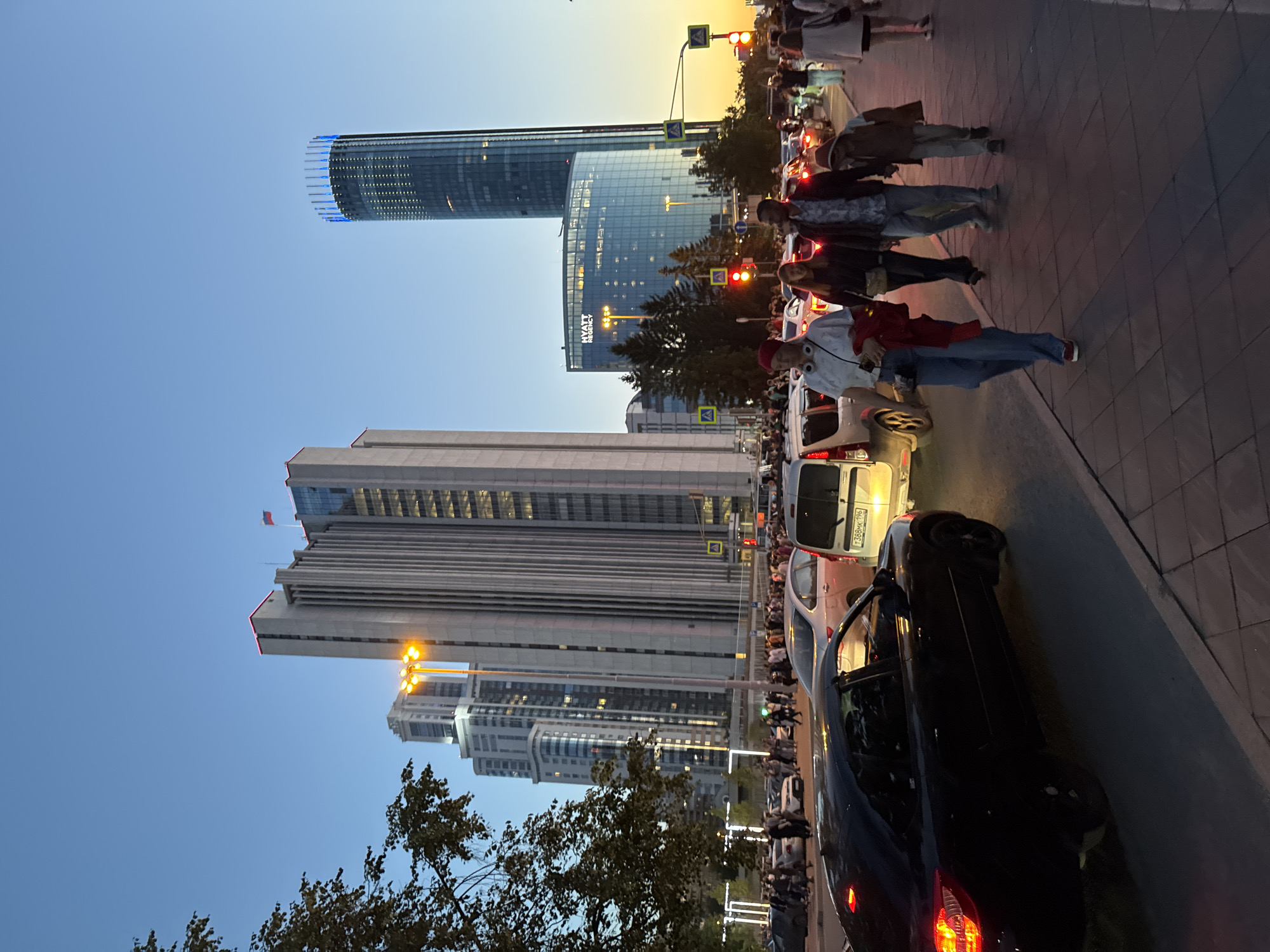 This image captures a street scene in Yekaterinburg, Russia, likely in the evening or dusk, given the lighting conditions. Dominating the background are modern high-rise buildings, including a distinctive cylindrical tower illuminated with blue lights and a larger, rectangular building with a repeating vertical design. To the left, another tall residential building with multiple windows is visible.
In the foreground, a street is filled with a significant amount of traffic, with cars stopped, suggesting congestion. A group of people are walking on the sidewalk, some in casual attire. Among them, a man in a patterned shirt and jeans is walking with a child. A woman with headphones and a red jacket draped over her shoulder is also prominent. The sidewalk is paved with square tiles. A black car is parked on the left side of the frame, and a silver van is visible in the traffic. Traffic lights are illuminated red. The sky is a clear gradient of blue transitioning to a warm yellow-orange near the horizon, indicative of sunset or twilight.