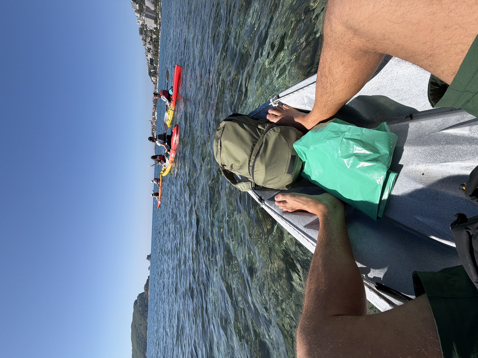 The image shows a first-person perspective from a kayak on the Adriatic Sea, near Budva, Montenegro. The water is clear, revealing the seabed. In the foreground is a kayak with a green backpack and a green plastic bag on board. Bare feet and legs, likely belonging to the person taking the photo, are visible. In the distance, a line of kayakers, wearing life vests, paddles along the coast. On the horizon, buildings in Budva are visible, including a mountainous region on the left. The sky is blue and cloudless.