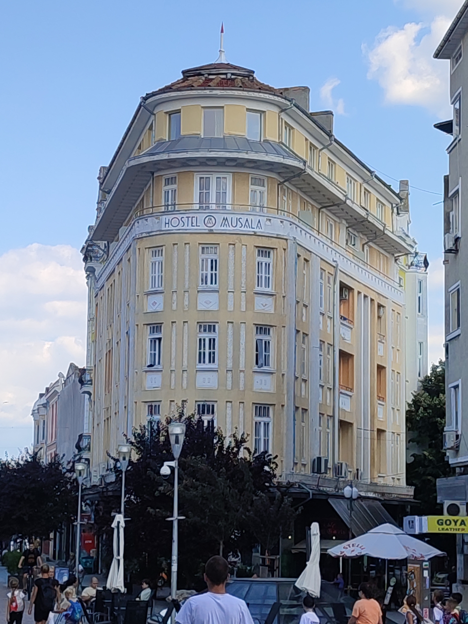 The image captures the Hostel Musala building, a prominent landmark in the city of Varna, Bulgaria, located on a bustling street. The building itself is a multi-story structure with a distinctive curved facade, painted in a pale yellow color with white architectural details. A circular turret with a red-tipped spire crowns the building. The hostel's name, "HOSTEL MUSALA," is visible in a stylized font on the facade.
Below the hostel, the street level is characterized by businesses, outdoor seating areas for cafes and restaurants, and streetlights. Several trees with dark foliage are planted along the sidewalk.
People are seen on the street, walking, cycling, and seated at outdoor tables. The overall atmosphere suggests a vibrant urban environment on a clear day, with some clouds present in the sky. The building's architecture is reminiscent of early 20th-century styles, possibly Art Nouveau or similar.
In the foreground, the backs of several people are visible, suggesting the photographer is observing the scene from a street-level perspective. To the right of the Hostel Musala, another multi-story building is partially visible, painted in a lighter gray. A yellow sign reading "GOYA LEATHER" is affixed to its facade. The presence of air conditioning units on some of the buildings indicates a warm climate.