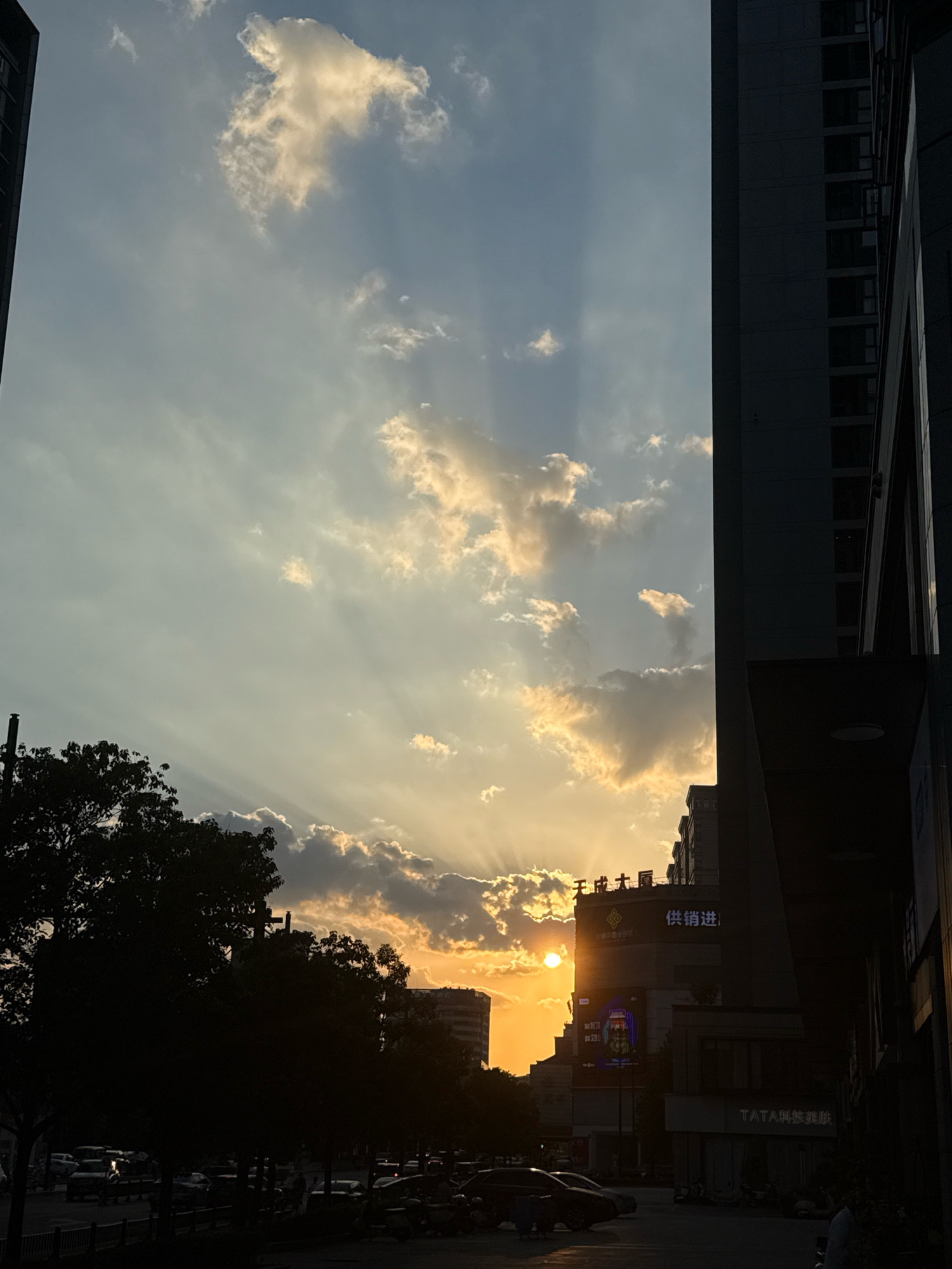 The image captures a sunset scene in Yiwu, China, viewed from a street level perspective between two tall buildings. The sky is filled with dramatic sun rays piercing through scattered clouds, illuminated by the setting sun which is partially obscured by a building and trees. The foreground features a silhouetted street with parked vehicles, including cars and motorcycles, and a few indistinct human figures. The buildings on either side are dark and imposing, with one on the right displaying visible signage. A large tree on the left also contributes to the silhouette effect, framing the sky. The overall impression is one of urban twilight with the natural spectacle of the sunset dominating the upper half of the frame.