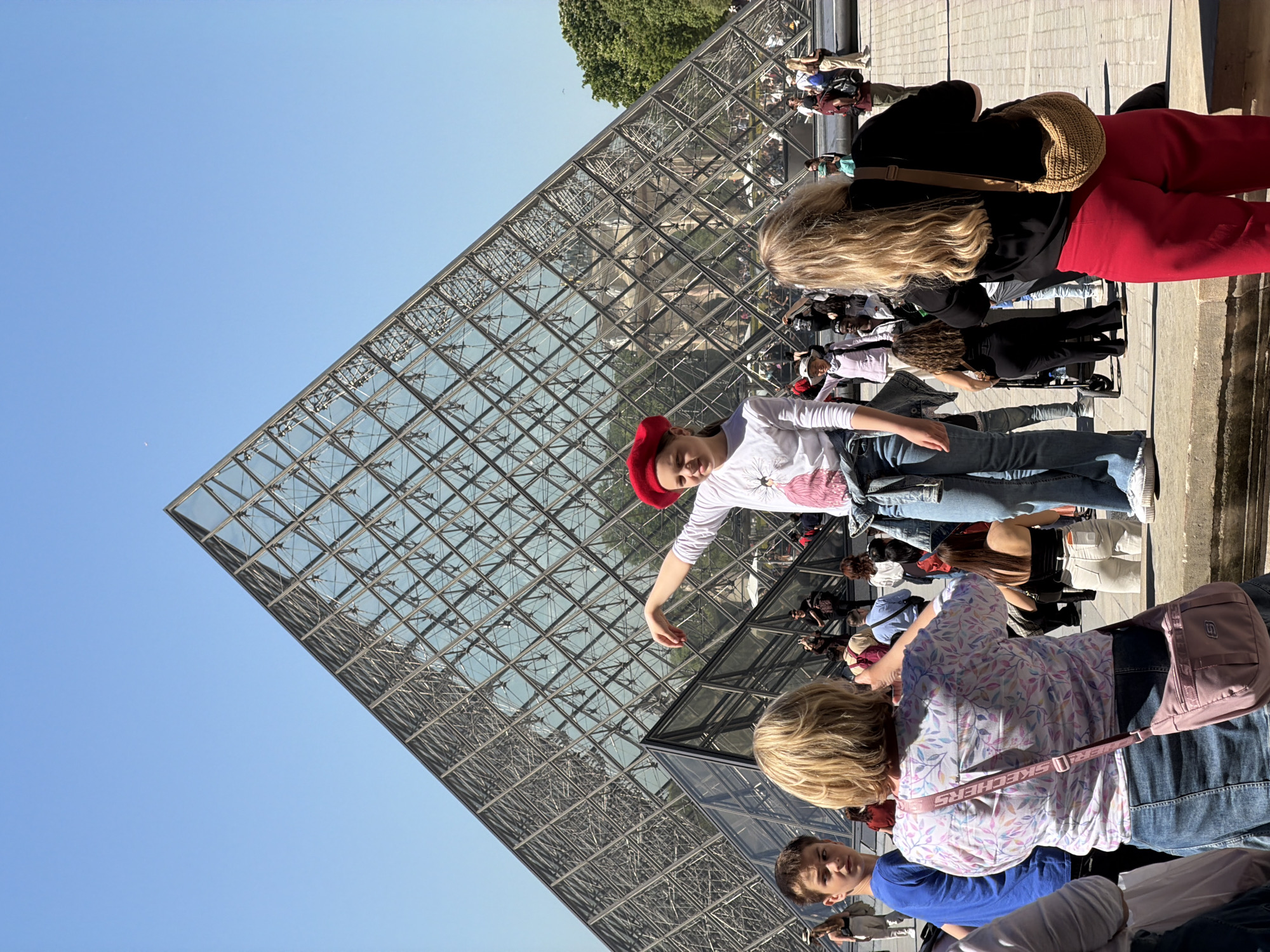 The image shows the Louvre Pyramid in Paris, France, with several people standing around it. A girl in a red beret and a white shirt with a floral design has her arm outstretched towards the pyramid. A woman with long blonde hair is wearing a black jacket and red pants, and carrying a woven bag. Another woman with blonde hair is wearing a light-colored floral print shirt and a pink Skechers bag. A young man in a blue shirt is also visible in the foreground. The pyramid's glass panels reflect the sky and other visitors.