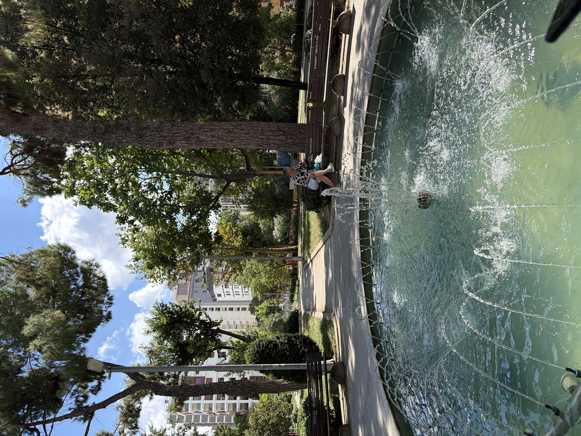 The image depicts a public park in Kadıköy, Turkey, featuring a circular fountain in the foreground with water jets spraying upwards. Beyond the fountain, a paved pathway winds through the park, flanked by benches and lush greenery. In the mid-ground, a person is seated on a park bench, looking down at a phone. Behind the park, tall apartment buildings rise against a backdrop of a blue sky with scattered clouds. The overall scene conveys a serene urban park setting on a sunny day.