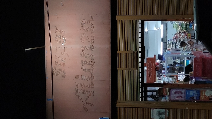 A store at night, illuminated from within, shows a person looking at shelves stocked with various goods. A closed, textured metal door to the left has illegible writing on it. The building's facade is wooden slats with metal security bars partially visible. The scene is dark, with most light emanating from the store itself.