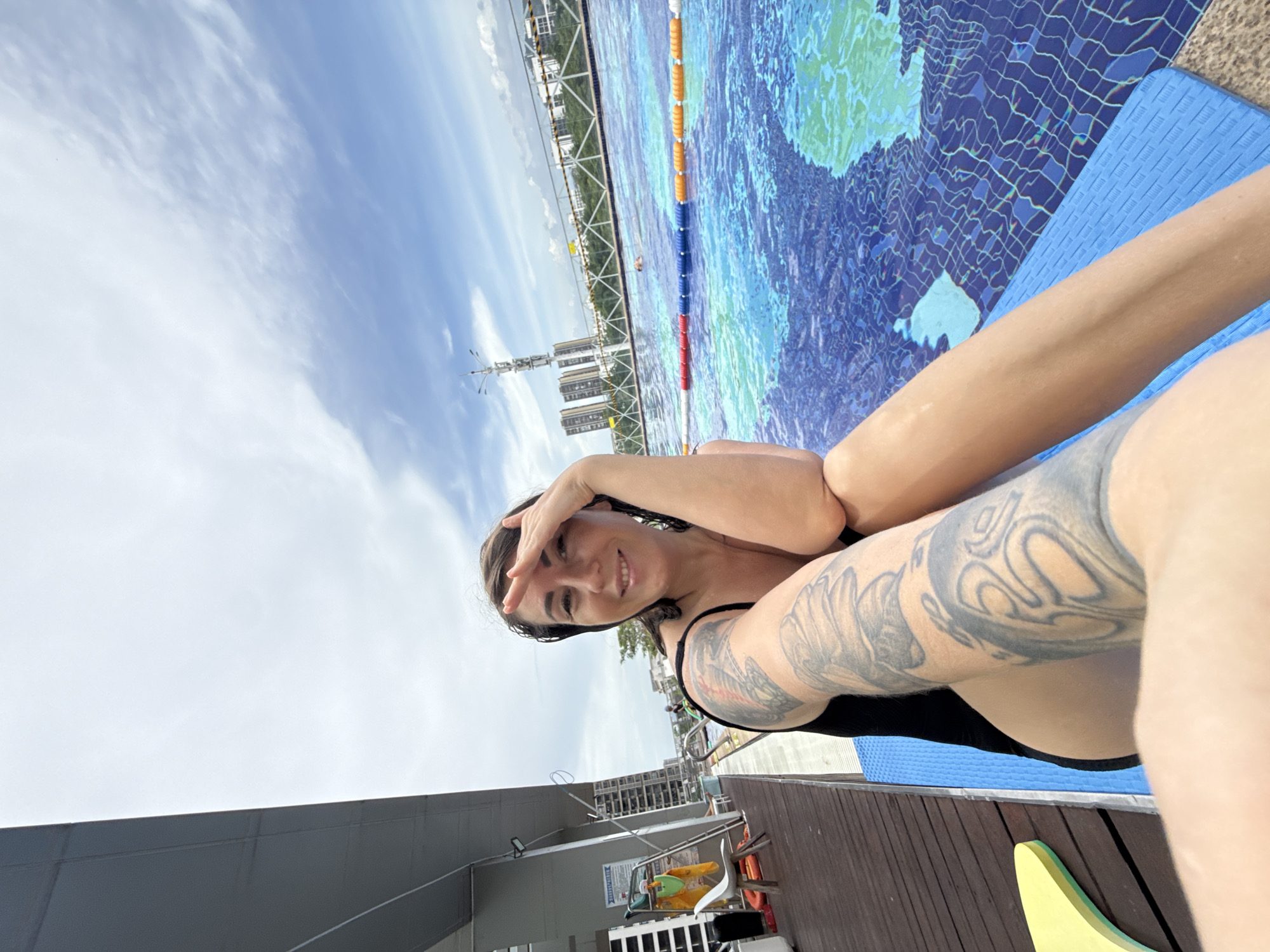 A woman with tattoos on her left arm is sitting by a swimming pool in Huizhou, China. She is wearing a black swimsuit and is shielding her eyes from the sun with her right hand. The swimming pool is blue with lane markers and has a view of city buildings and a construction crane in the distance. The sky is partly cloudy. To the left of the woman is a wooden deck with chairs and a yellow flotation device.