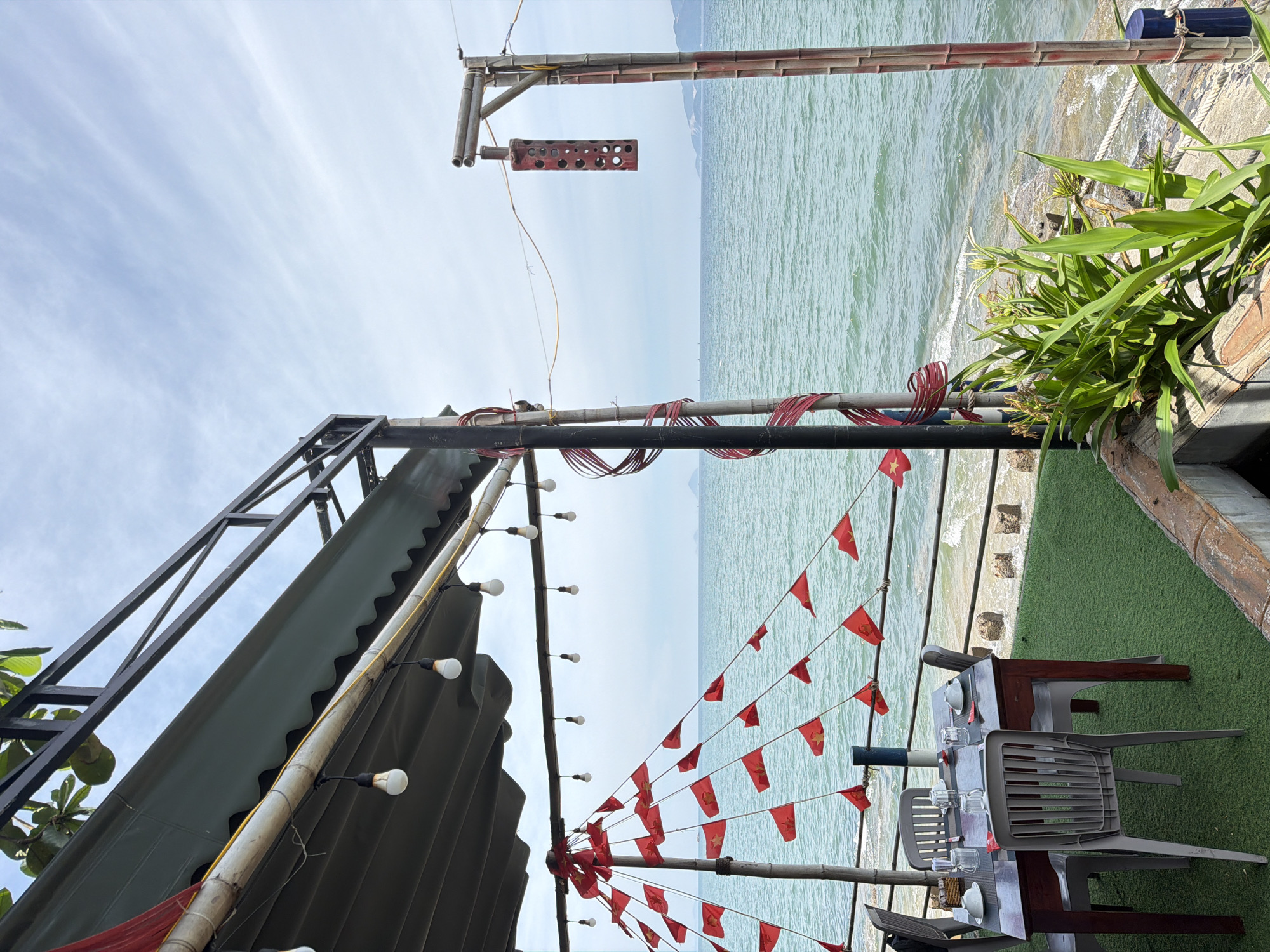 The image depicts an outdoor dining area by the sea in Nha Trang Ward, Vietnam. A table is set with white plates and glasses for a meal, positioned on a green, artificial turf surface. The area is decorated with numerous small red flags, strung along wires. Overhead, a green awning is supported by a metal frame. A metal pole with decorative red coils stands to the left, while another bamboo pole with red markings supports a hanging lantern on the right. The sea is visible in the background, with a calm, blue-green color. The sky is partly cloudy. In the foreground, lush green plants are growing. The overall scene suggests a casual, seaside restaurant or cafe.