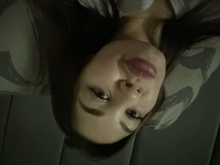 The image displays a close-up portrait of a person, seemingly a young woman, captured in a low-light indoor setting. The image is rotated approximately 90 degrees counter-clockwise, showing the person's head tilted to the right.
If rotated to an upright orientation, the woman's face would be central, looking directly towards the camera with a soft, calm expression. She has dark hair that frames her face. Her eyes are dark, possibly brown, with visible eyelashes that suggest the use of mascara. Her eyebrows are dark and well-defined. Her lips are full and prominent, accentuated with a glossy, rosy-pink or light red lipstick. Her skin appears smooth and has a natural complexion, with subtle highlights on her nose and cheeks.
She appears to be lying down, with her head resting on a light-colored pillow that has a subtle, abstract or camouflage-like pattern in shades of gray and white. A light-colored garment, possibly a shirt or top, is visible around her neck and upper chest area. The overall lighting is dim, suggesting a relaxed evening or nighttime atmosphere. The background is dark and indistinct, focusing attention primarily on her face. No text is visible in the image.
