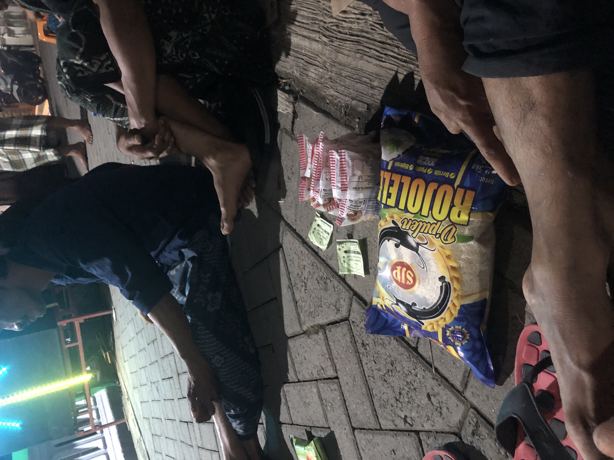The image depicts a street scene in Tenggilis Rejo, Indonesia, with several individuals present. In the foreground, a bag of "Rojolele" rice, weighing 2.5kg, is prominently displayed on a paved surface. Scattered around the bag are several small packets of what appear to be snacks or seasonings, along with two Indonesian Rupiah banknotes. To the left, a pair of red and black flip-flops lies on the ground.
Two individuals are partially visible, seated on the ground. One person, wearing a dark blue collared shirt and patterned pants, is seen from the side, with their legs extended. Another individual, in the lower right of the frame, is also seated, with their arm and hand resting near the rice bag. Their legs are visible, and they appear to be wearing dark clothing. In the background, another person is seen standing, wearing a checkered sarong. The environment suggests an outdoor setting at night or in low light conditions, illuminated by artificial lighting, including a bright green and yellow light source in the upper left. The ground is paved with rectangular blocks. A motorcycle is visible in the background, along with a small orange stool.