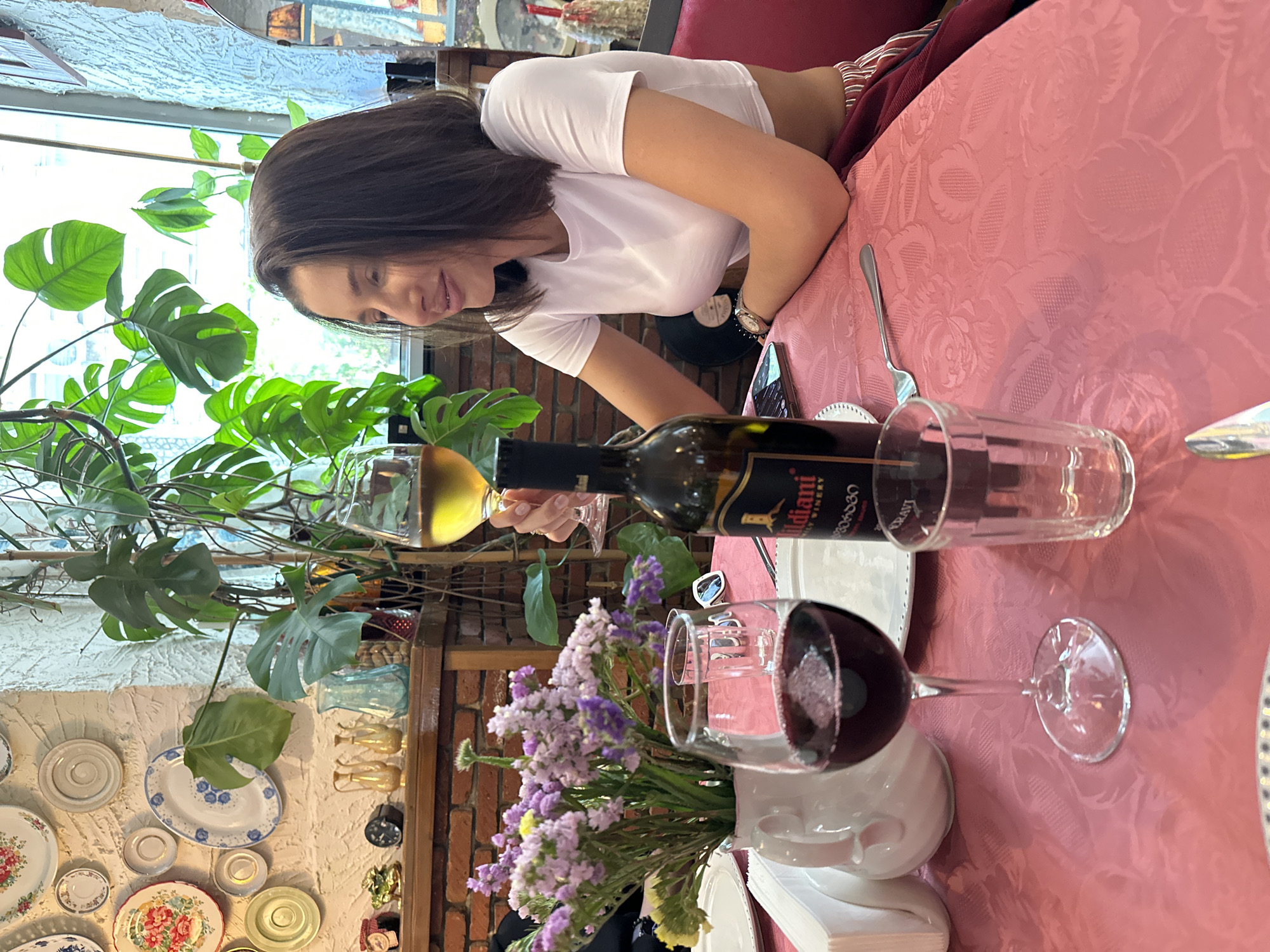 A woman in a white t-shirt is seated at a table in a restaurant in Tbilisi, Georgia. A bottle of Tbilvino Mildiani wine, a white wine glass, and a red wine glass are on the table. Purple flowers are arranged in a vase. A fork and a side plate are visible. The table is covered with a pink tablecloth. Behind the woman, indoor plants are present. Decorative plates are displayed on the wall. The scene is captured in daylight.
